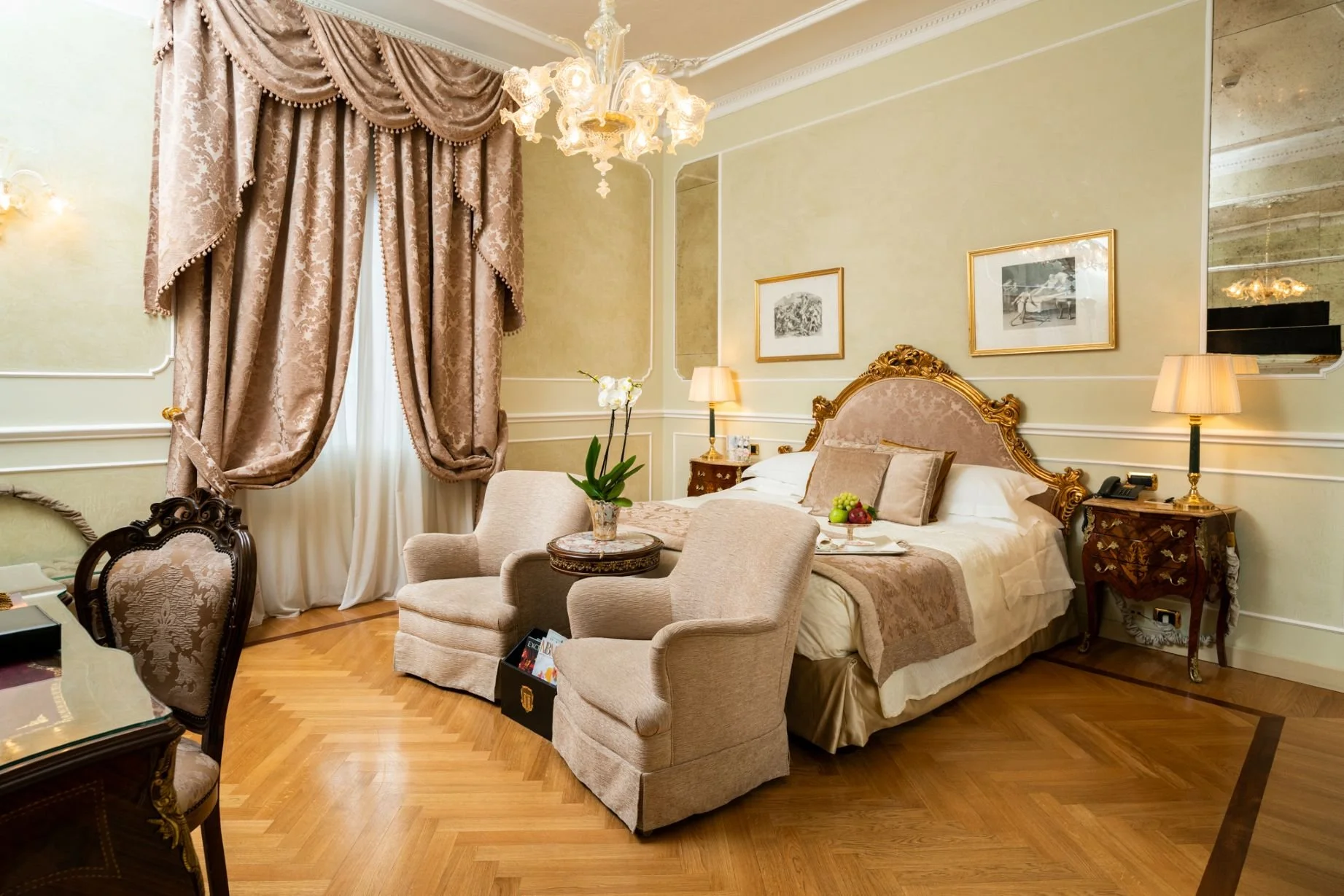 Elegant hotel room with a large bed, ornate gold headboard, beige and gold decor, floral curtains, sitting area with two armchairs and a small table, lamps, framed artwork, and hardwood floors.