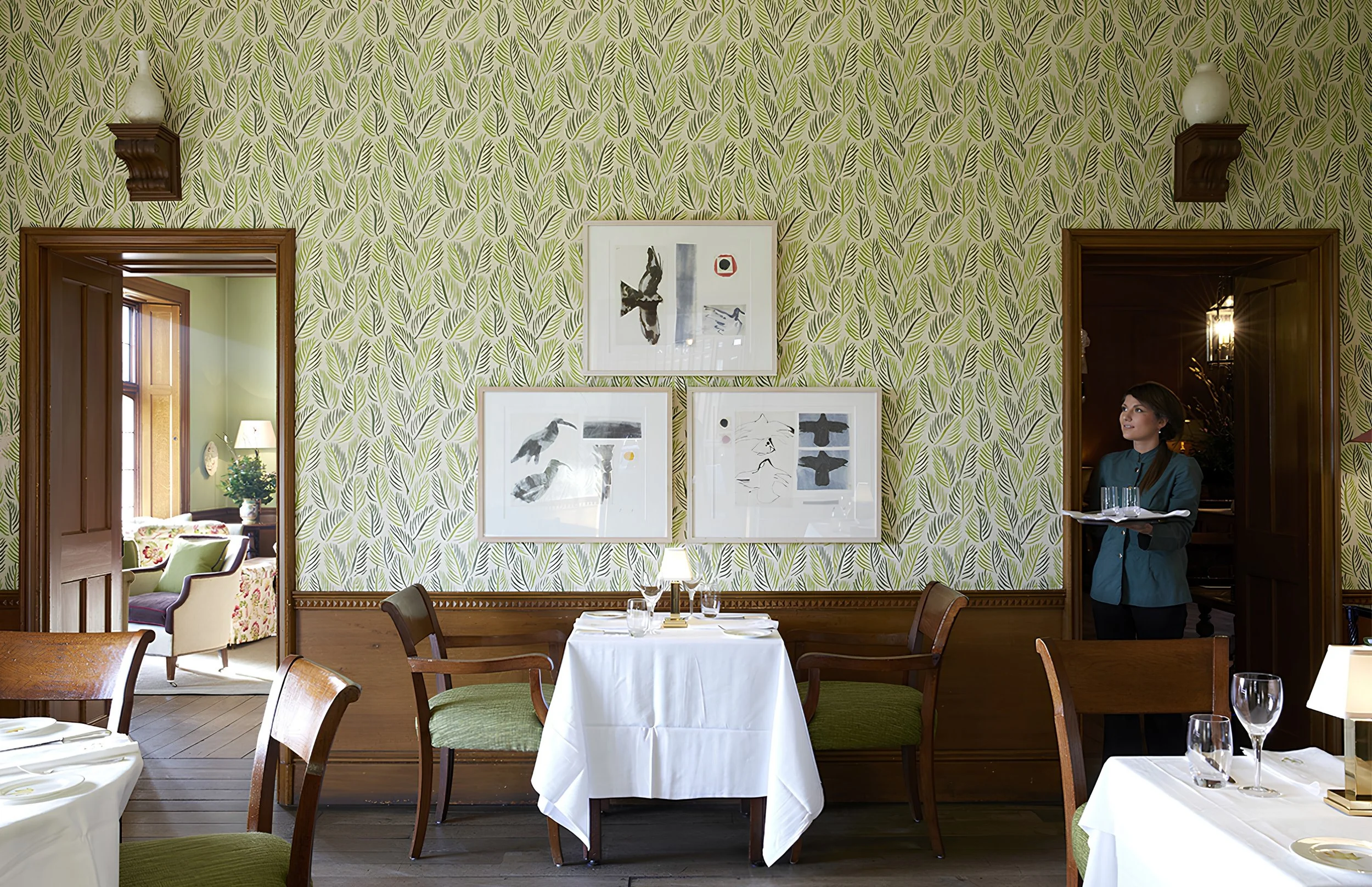 A dining room with green patterned wallpaper, framed artwork on the wall, and a woman in a blue shirt serving drinks. There are tables set for dining with white tablecloths, wine glasses, and silverware. A doorway reveals a windowed sitting area with