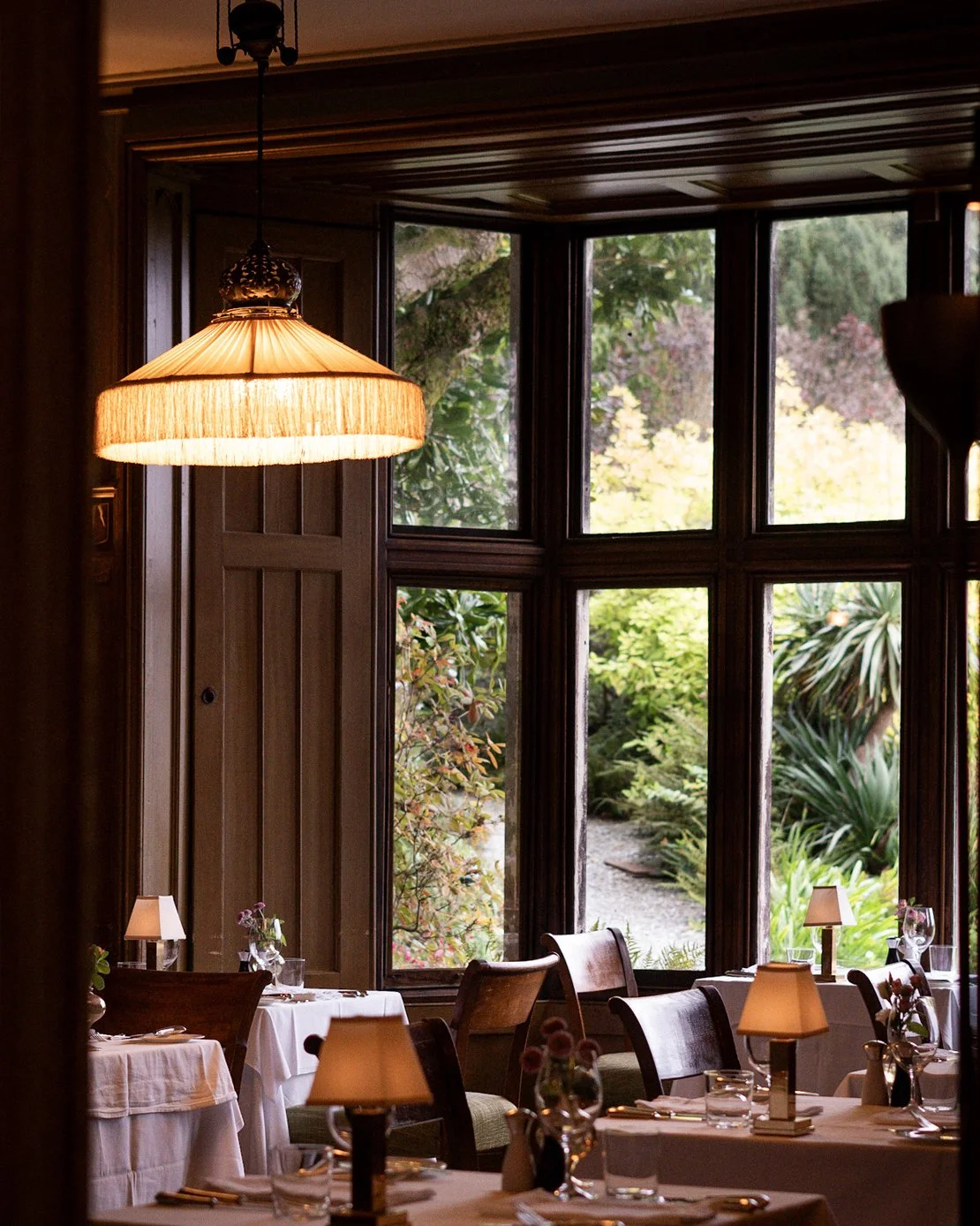 Interior of a cozy restaurant with tables set for dining, large windows revealing greenery outside, and warm lighting from table lamps and a hanging ceiling lamp.