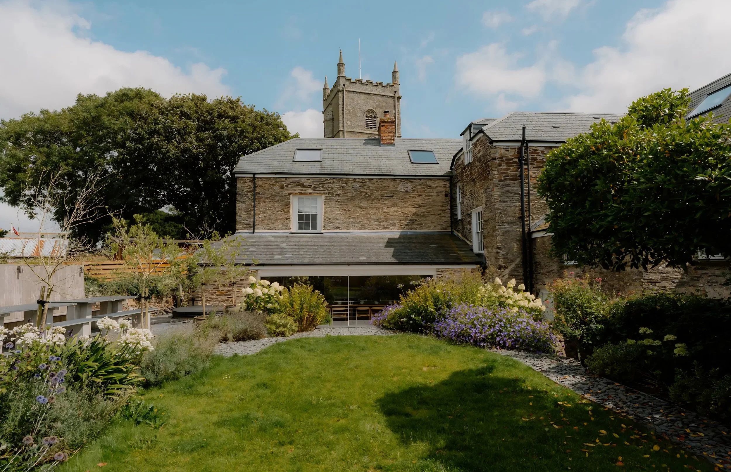 A garden with various plants and flowers in front of a stone house with a slate roof and skylights, a stone church tower with battlements and pointed turrets in the background, and a partly cloudy sky.