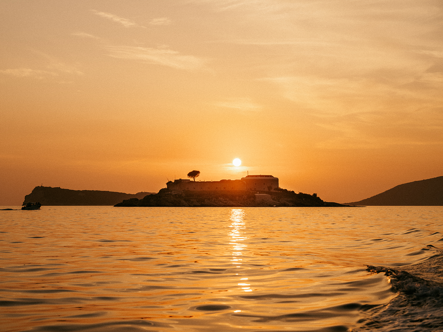 Sunset over an island with a small building and a tree, reflected in the calm water, with a boat on the water to the left.