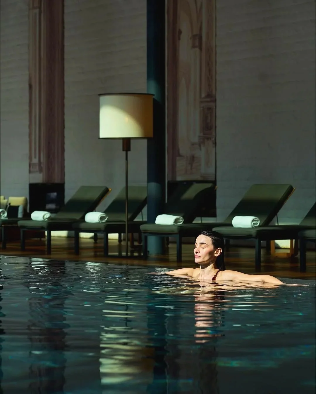 A woman relaxing in an indoor pool, with lounge chairs and a tall lamp in the background.
