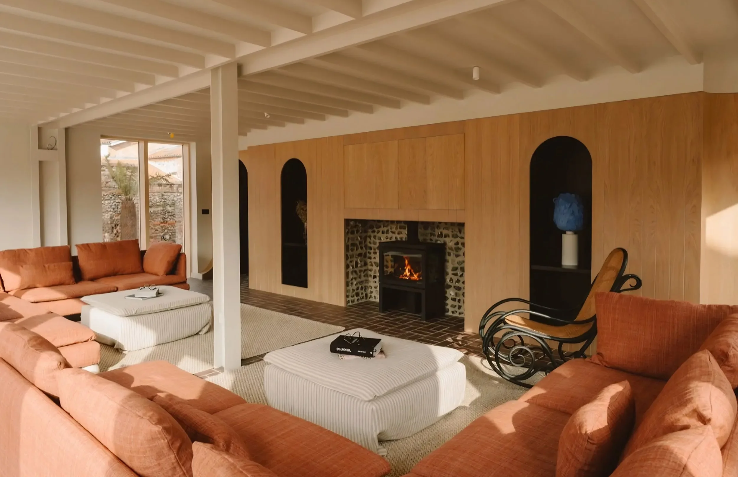Living room with a large pink sofa, white ottomans, a fireplace, wood paneling, and a wooden ceiling with exposed beams.