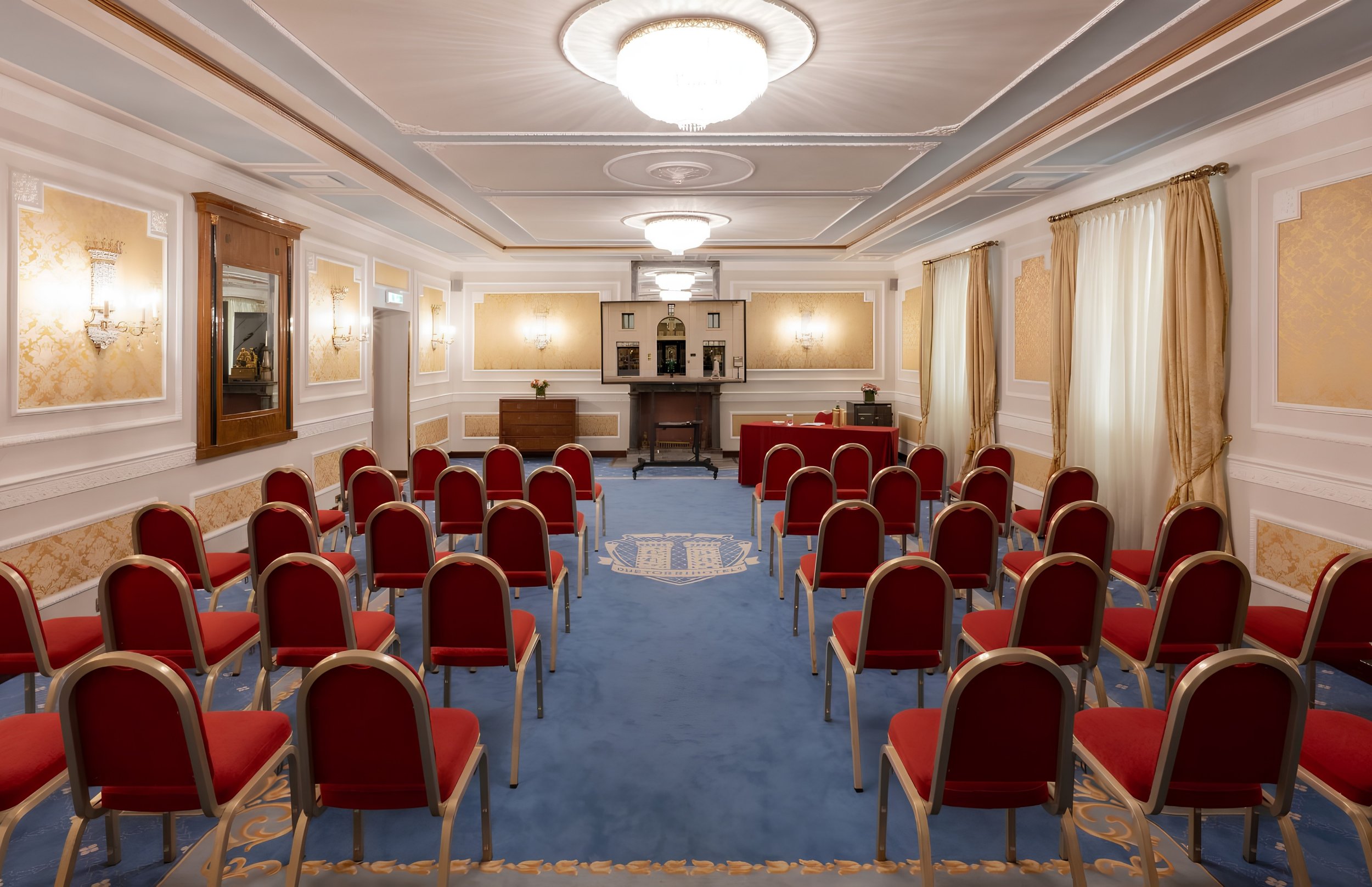 An elegant banquet room with rows of red cushioned chairs facing a small stage area with a table and flowers. The room features gold and white decor, chandeliers, and tall windows with gold curtains, creating a luxurious atmosphere.