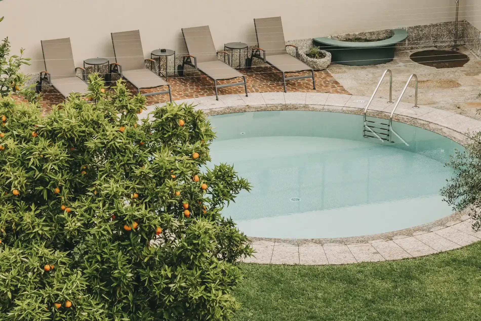An outdoor swimming pool with a metal ladder, surrounded by green grass, bushes, orange trees, and lounge chairs with small tables nearby. There are two small hot tubs or spas in the background.