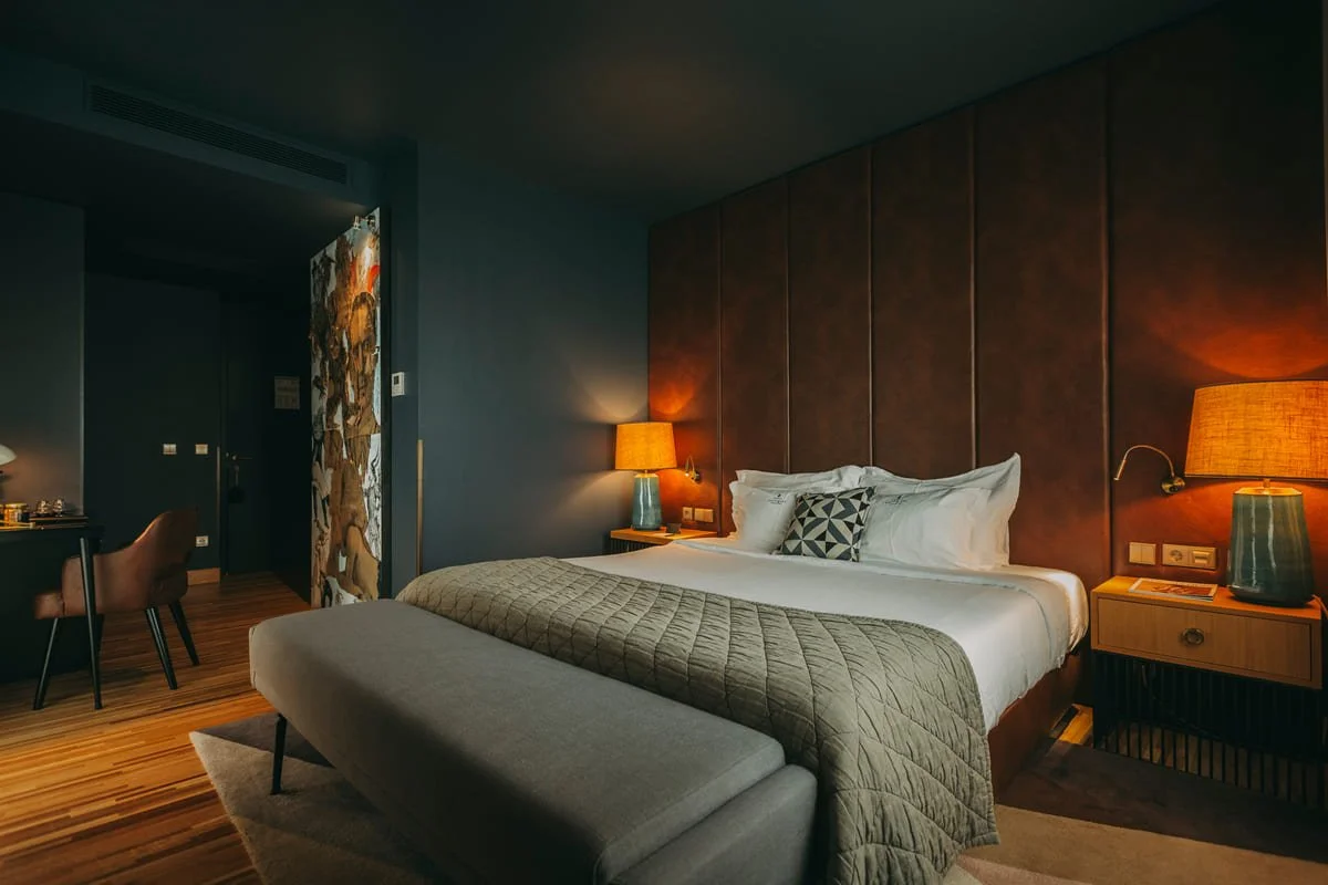 A cozy hotel bedroom with a large bed, dark brown padded headboard, two bedside lamps emitting warm light, a blue bedside table with a book, and a gray quilted blanket at the foot of the bed. There is a small seating area and a colorful curtain separating the room from the entrance.