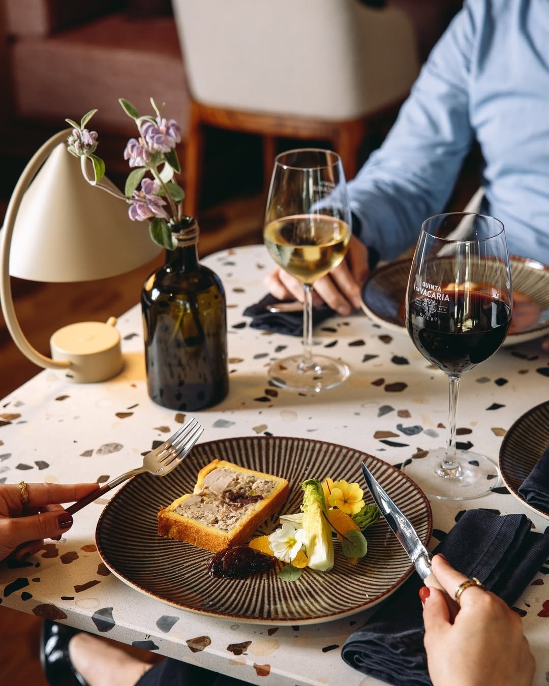 Table setting with a dessert plate, wine glasses with white and red wine, and a decorative vase with purple flowers.