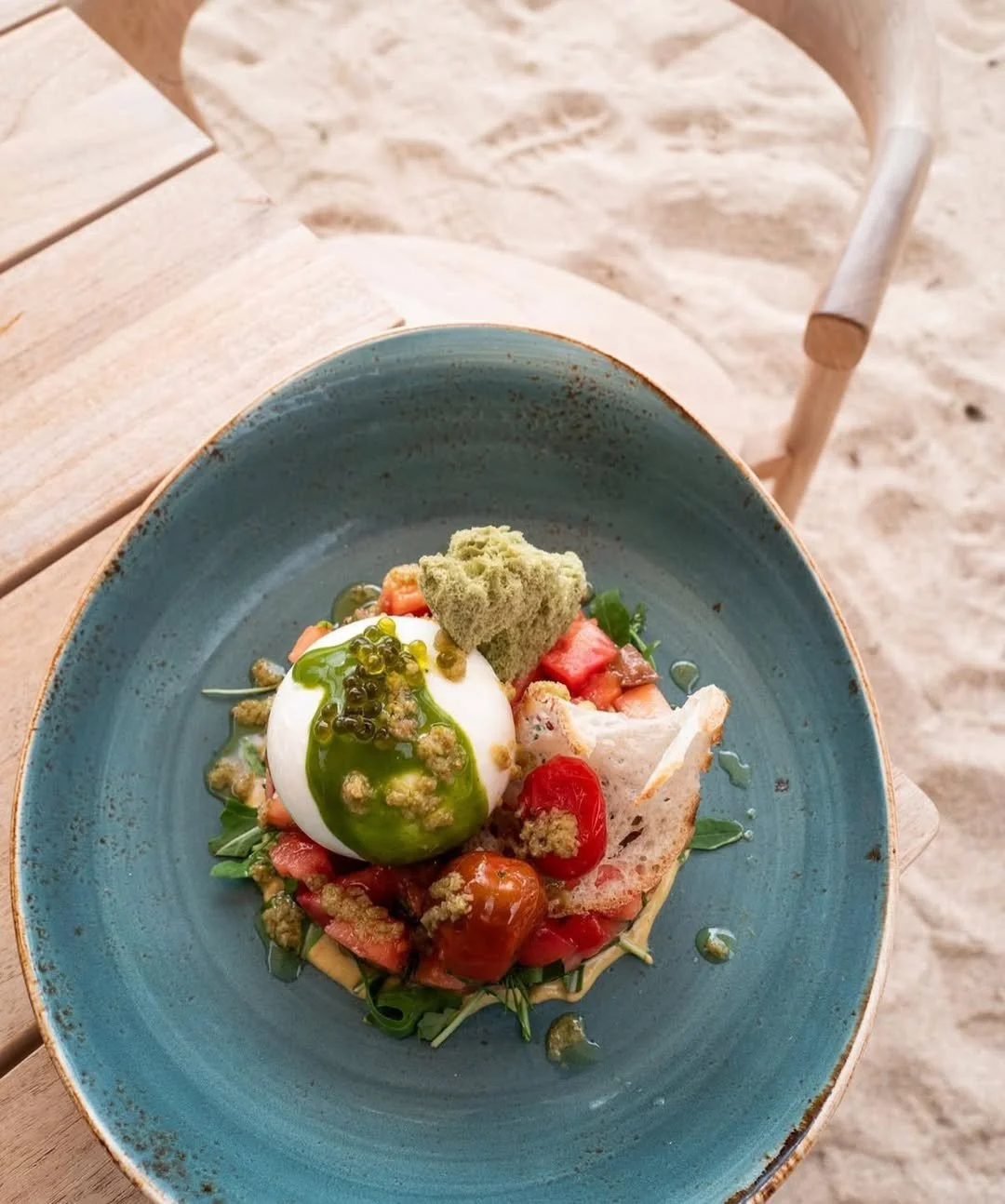 A plated dish with a poached egg topped with green sauce, cherry tomatoes, bread, and green herbs, served on a blue ceramic plate on a wooden table outdoors.
