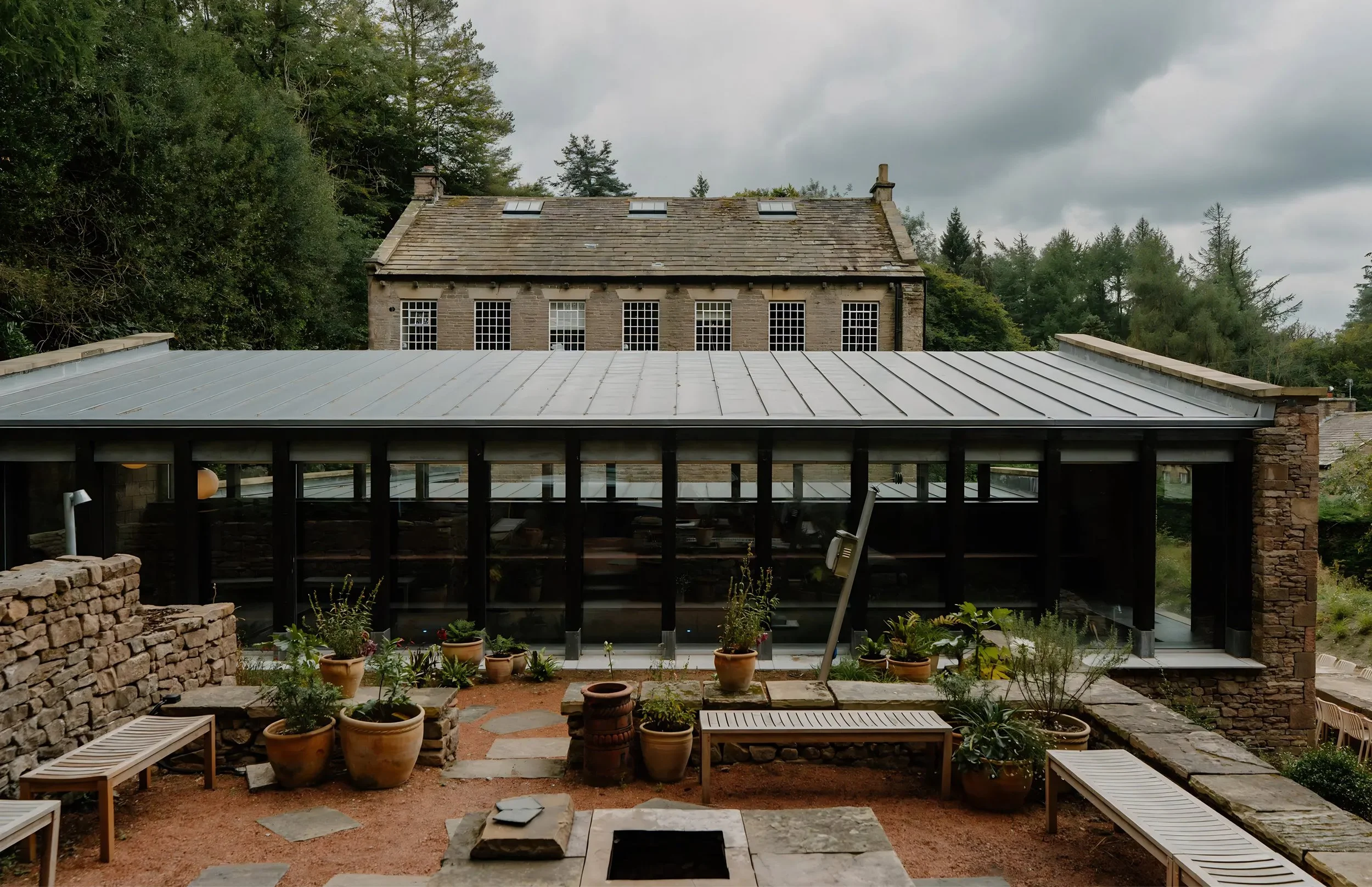 A view of a stone house with skylights on the roof, surrounded by trees under a cloudy sky. In the foreground, there is a patio with potted plants, benches, and a fire pit, with a modern metal roof covering part of the outdoor space.