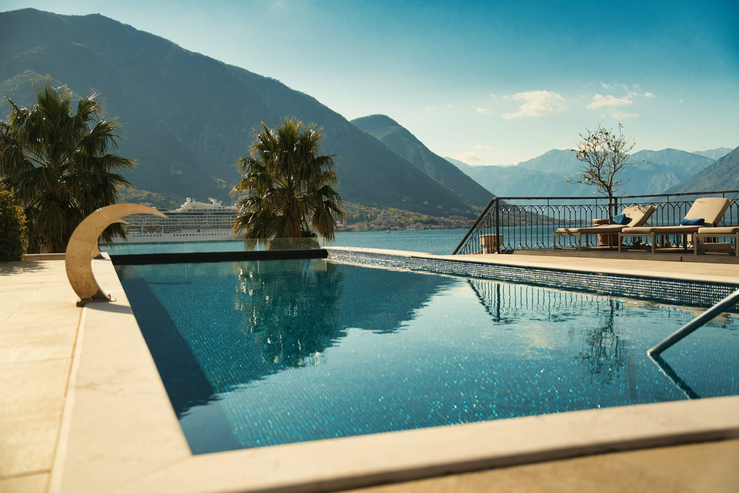 A swimming pool overlooking a lake with mountains in the background, lounge chairs by the poolside, and a cruise ship in the water.
