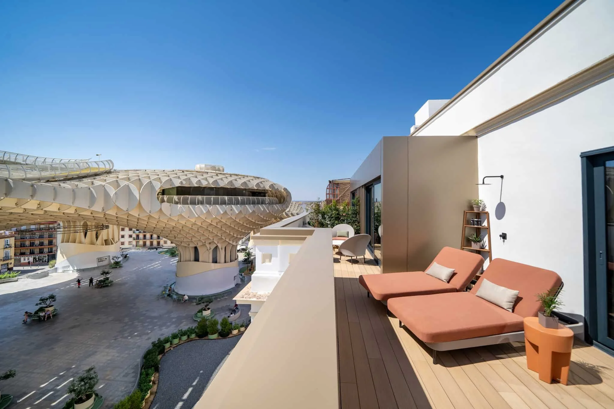 View from a balcony showing modern furniture, including orange lounge chairs and a small table, with a cityscape and distinctive architectural structure in the background under a clear blue sky.