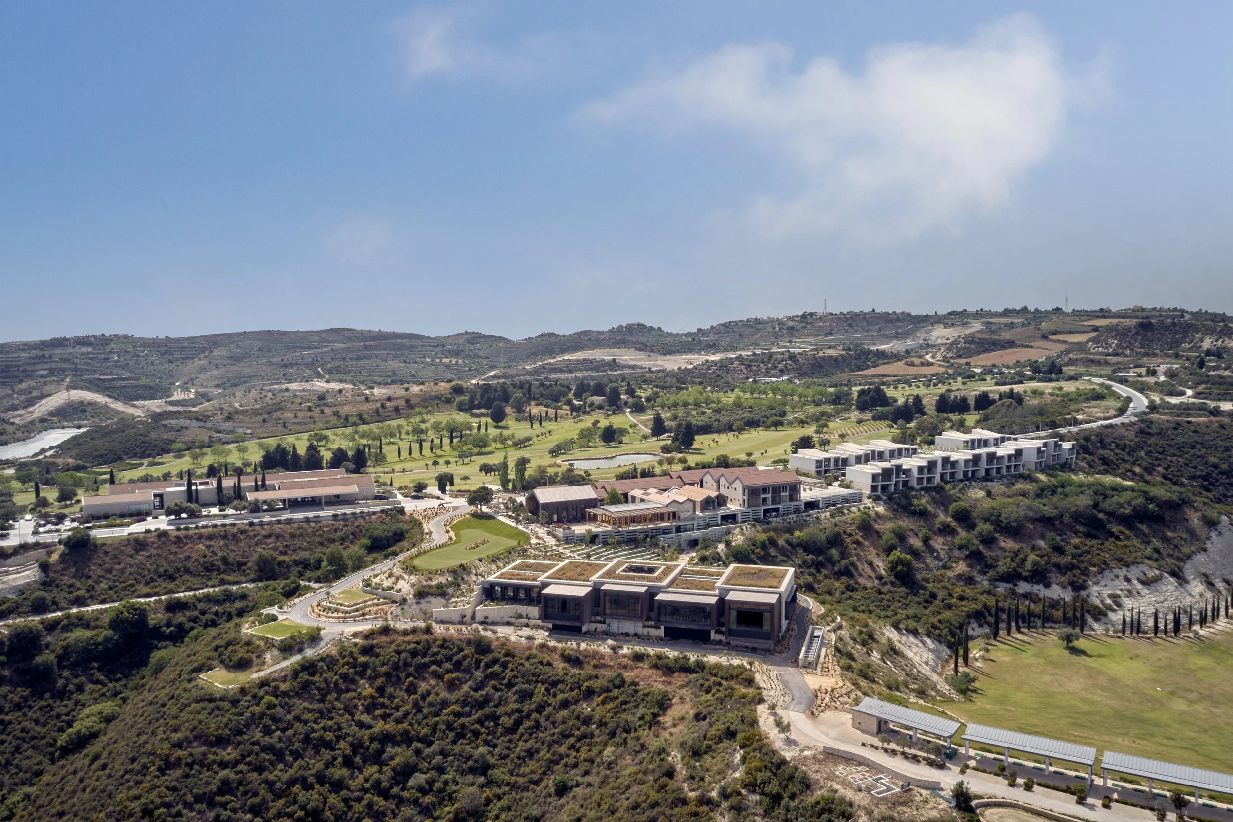 Aerial view of a hillside community with modern apartment buildings, lush green golf course, and rolling hills under a partly cloudy sky.