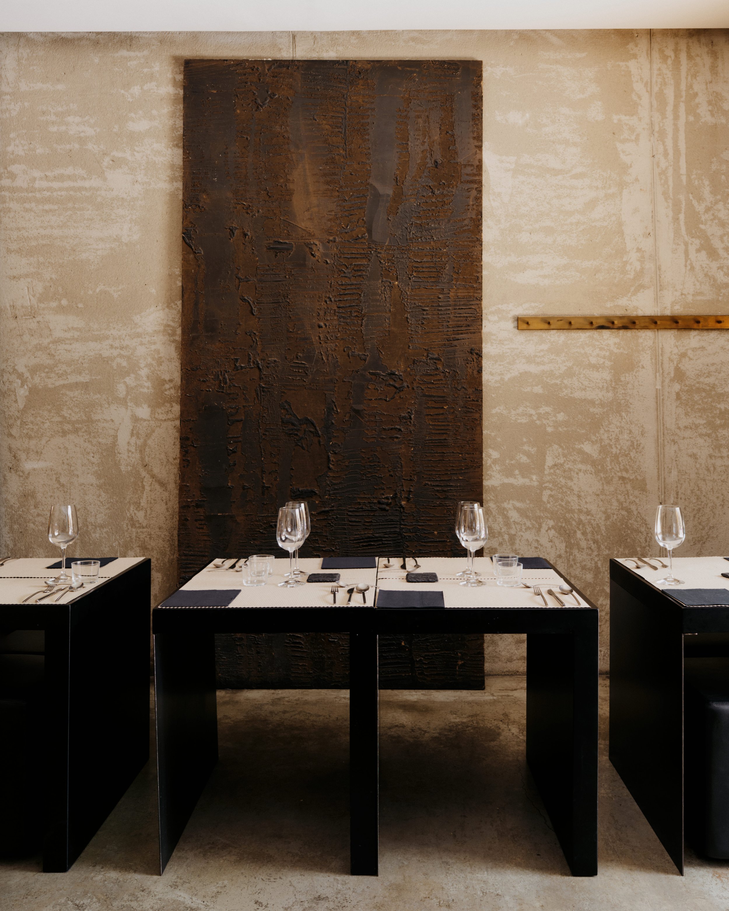 A dining table set for four with black chairs, white tablecloth, black placemats, wine glasses, and silverware, in front of a large textured brown abstract artwork on a beige wall in a modern restaurant or dining area.