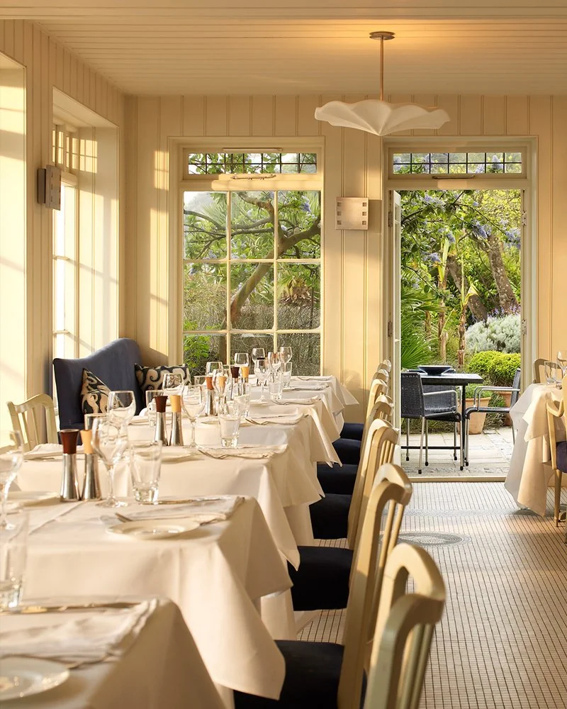 A cozy dining room with a long table set for a meal, featuring white tablecloths, wine glasses, silverware, and napkins. Large windows let in natural light and reveal a lush garden outside, with green trees and plants. There is a black bench with patterned cushions along the wall and a door leading to an outdoor patio.