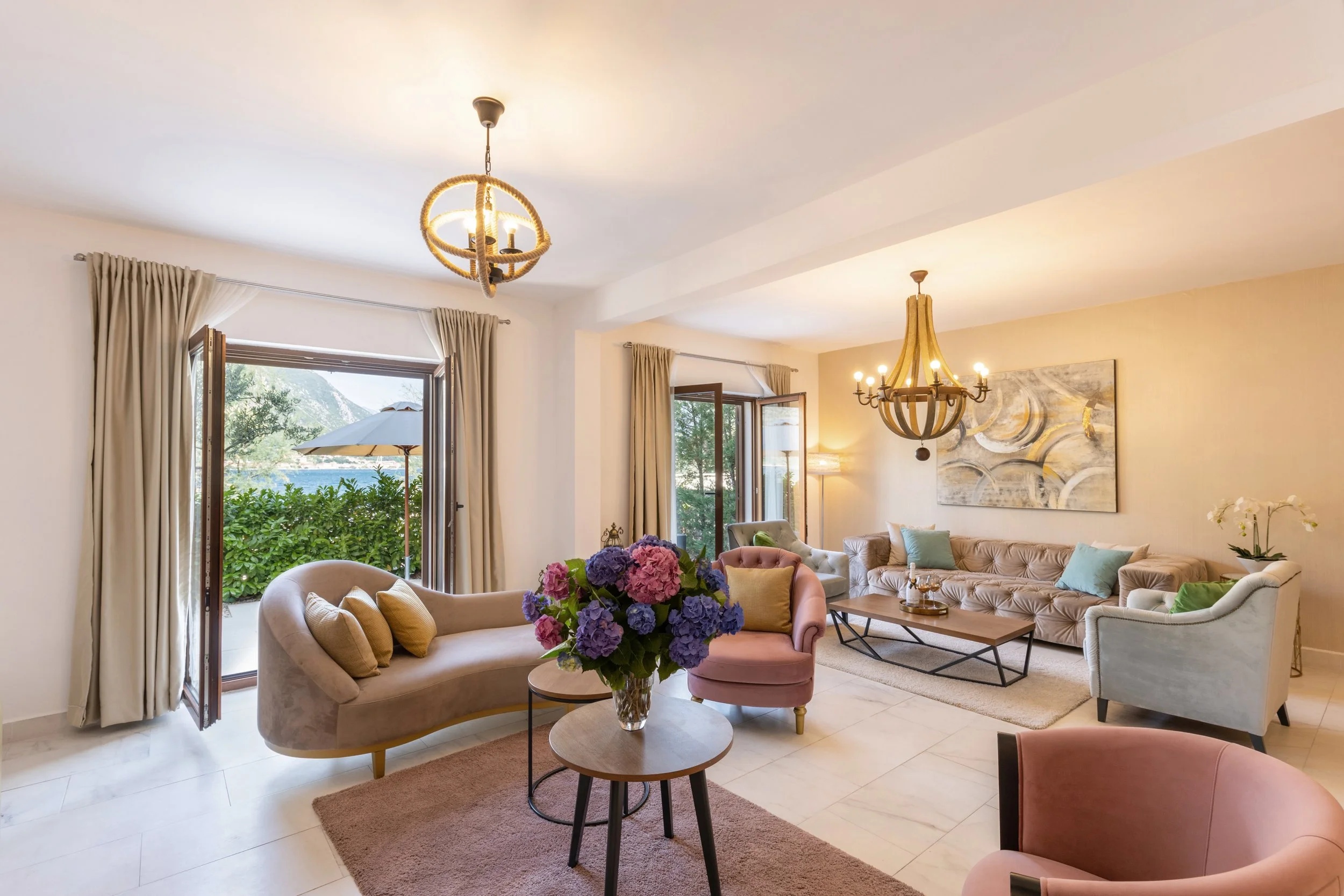 Living room with beige sofas, pink armchair, wooden coffee table, large painting on wall, chandelier, open windows with curtains, view of outdoor greenery and umbrella