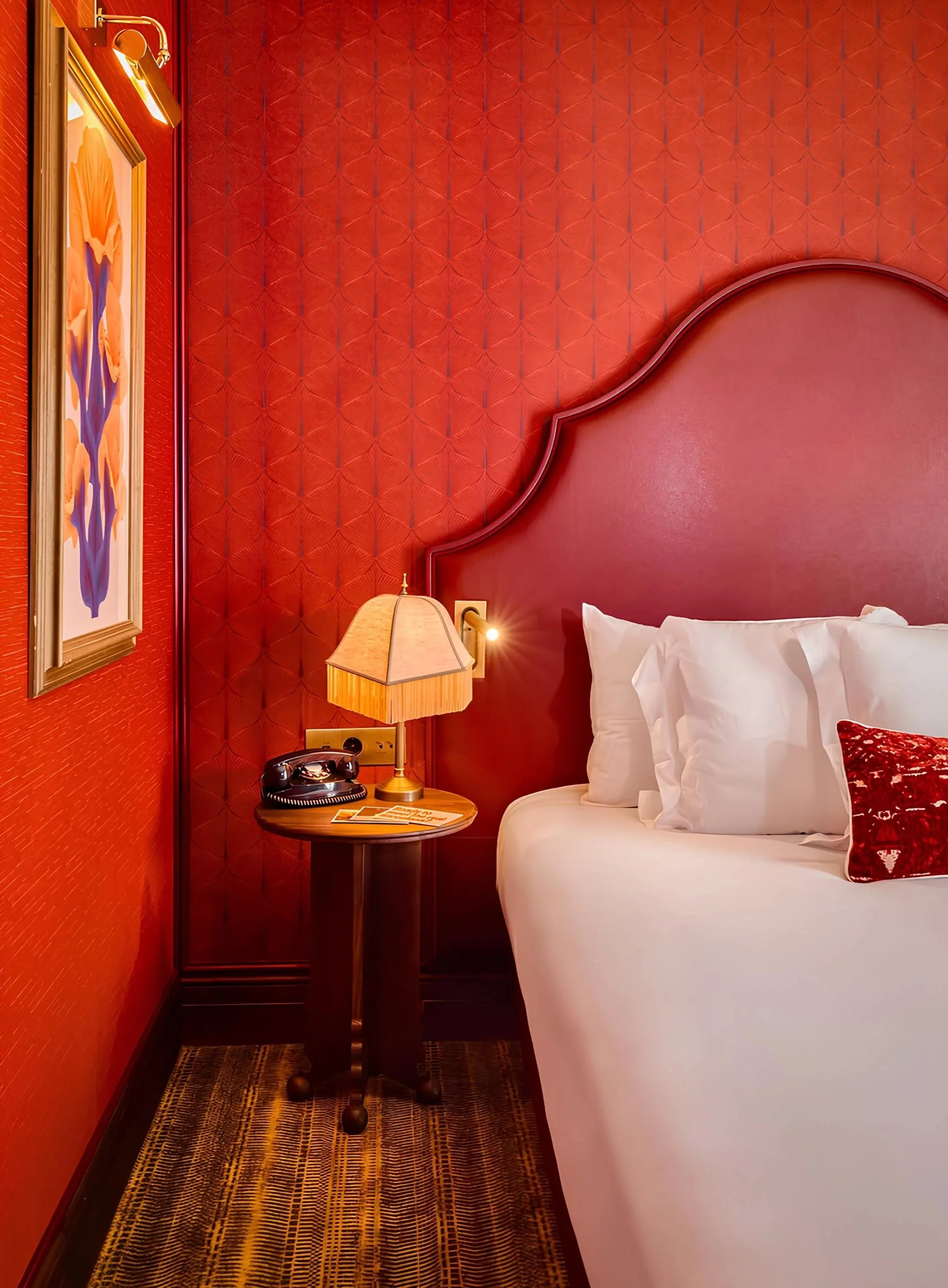 Close-up of a hotel bedroom with a white bed, a red decorative pillow, a wooden nightstand with a vintage black telephone, a table lamp with a beige lampshade, and a textured red wall with an abstract painting and a gold-colored picture frame. The side table, lamp, and part of the bed are visible, with a patterned carpet on the floor.
