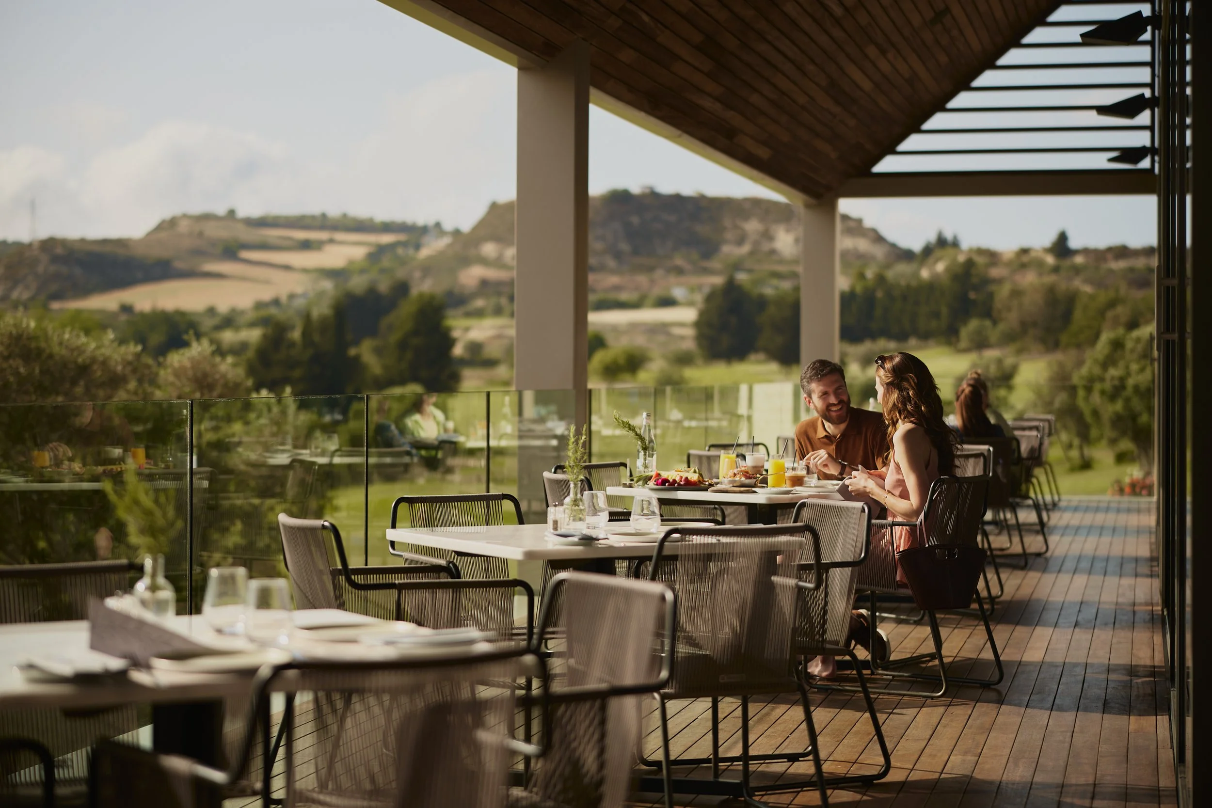 A couple sitting at an outdoor dining table with breakfast or brunch, smiling and talking, with a scenic hillside landscape in the background.
