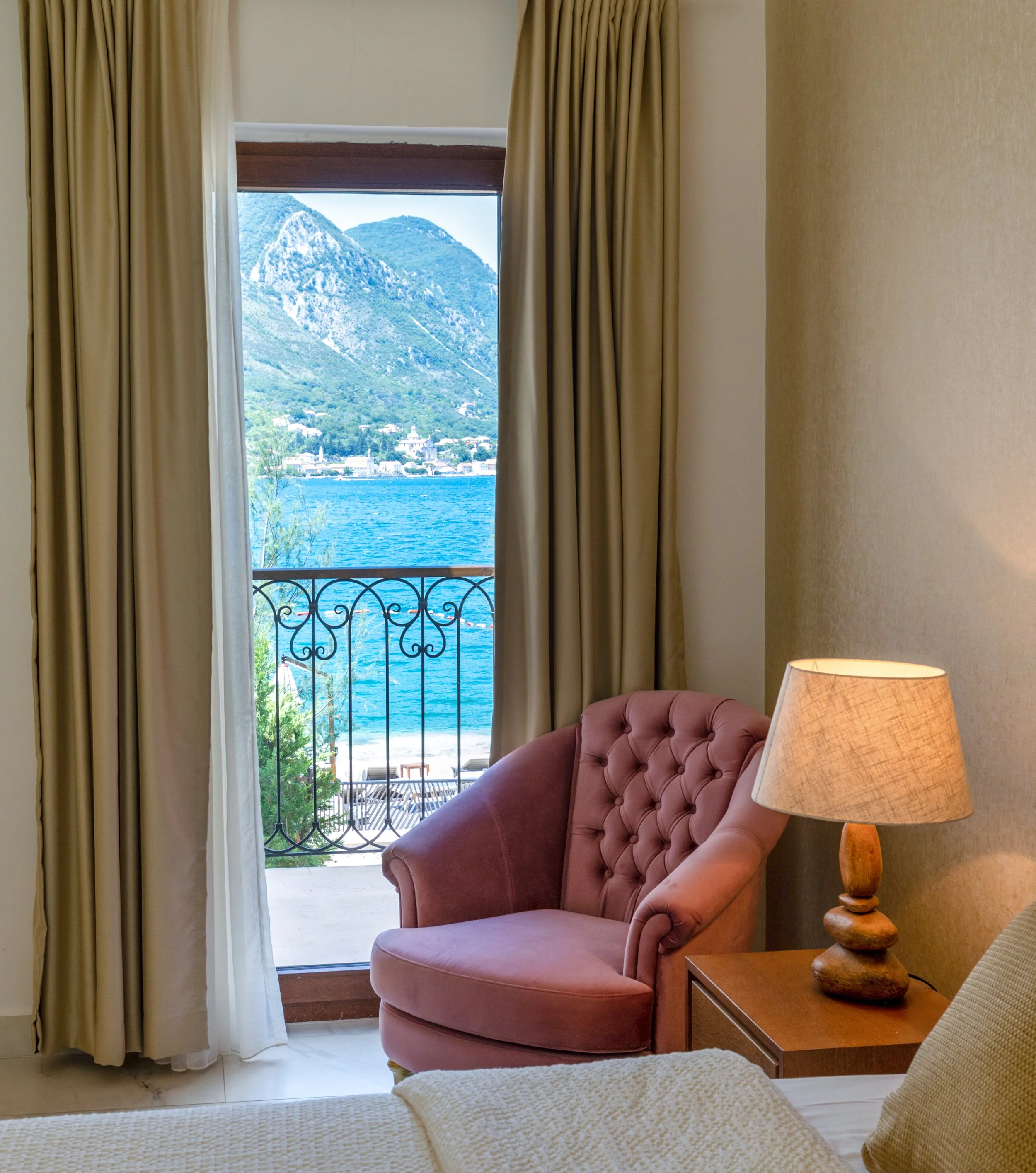 A room with beige walls, a pink upholstered chair, a wooden side table with a carved lamp, and a view of mountains, water, and a town through a window with beige curtains.