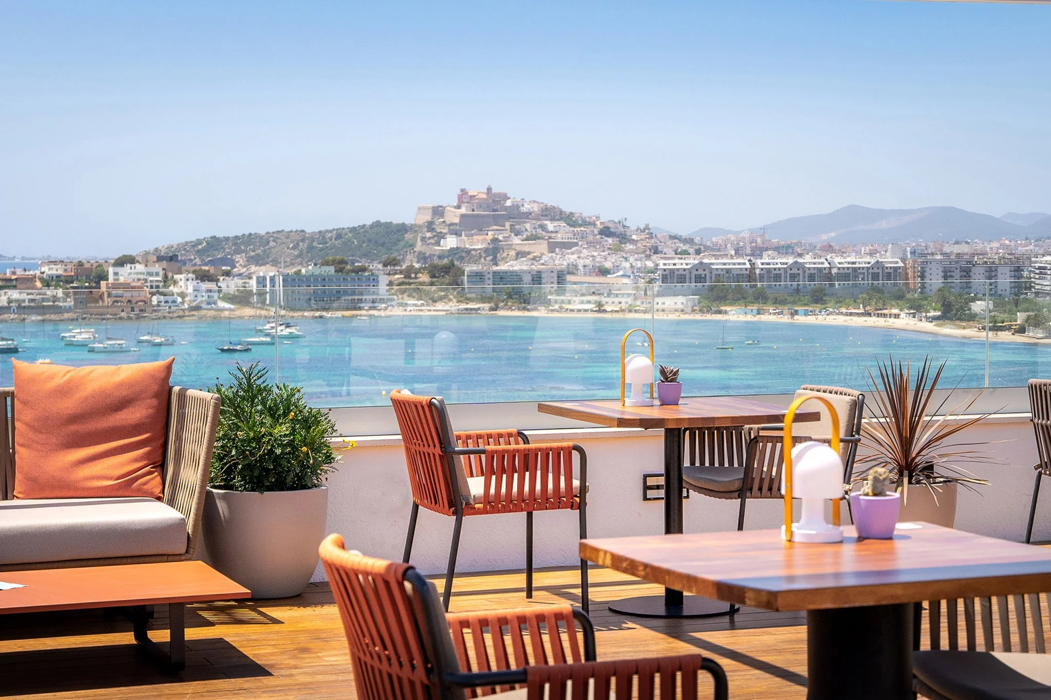 Outdoor terrace with seating overlooking a bay with sailboats and a cityscape with a hilltop fortress in the background.