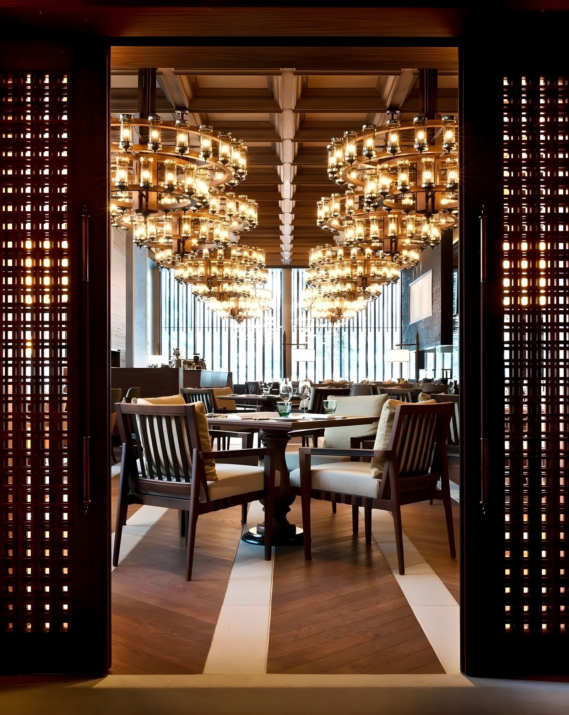 Elegant restaurant interior with chandeliers, wooden ceiling, and dining tables set with glassware viewed through an open doorway with decorative wooden panels.
