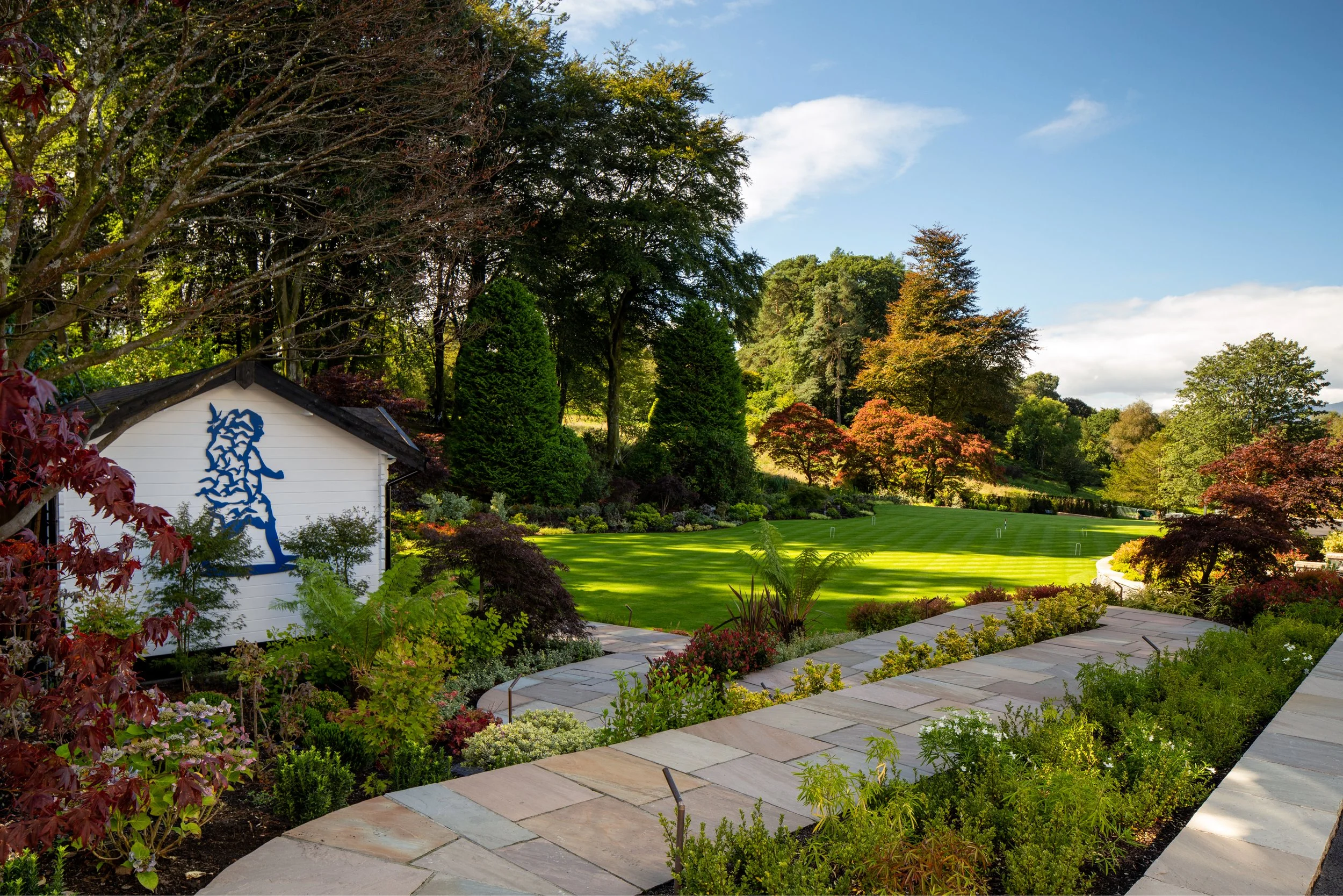 Beautiful landscaped garden with lush green trees, well-maintained lawn, flowering bushes, and a small white building with a blue horse sculpture on the side. Clear blue sky with a few clouds.