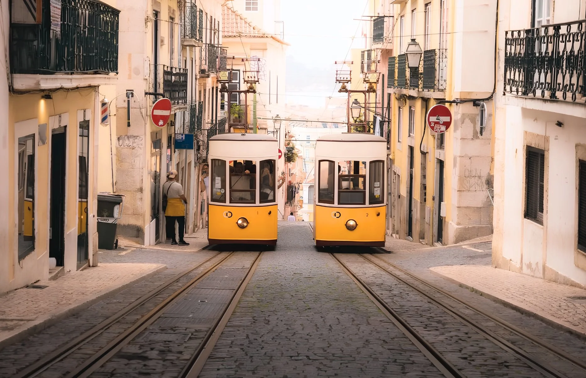 Two yellow streetcars traveling down narrow cobblestone street in a European city, flanked by multi-story buildings with balconies and black iron railings.