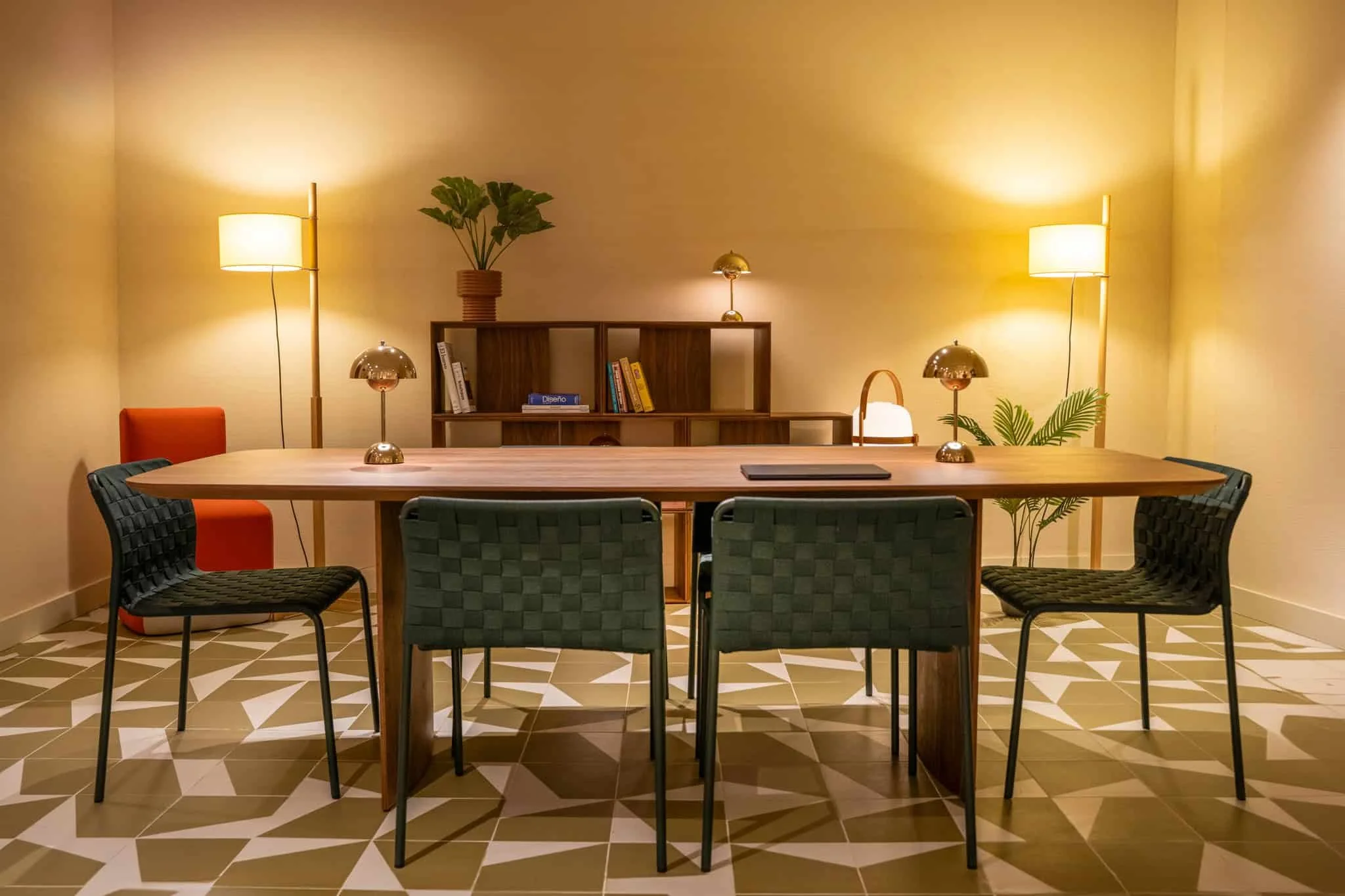 A dining or conference room with a large wooden table, six black woven chairs, two standing lamps, two table lamps, a bookshelf with books, a potted plant, and decorative lighting on a patterned tile floor.
