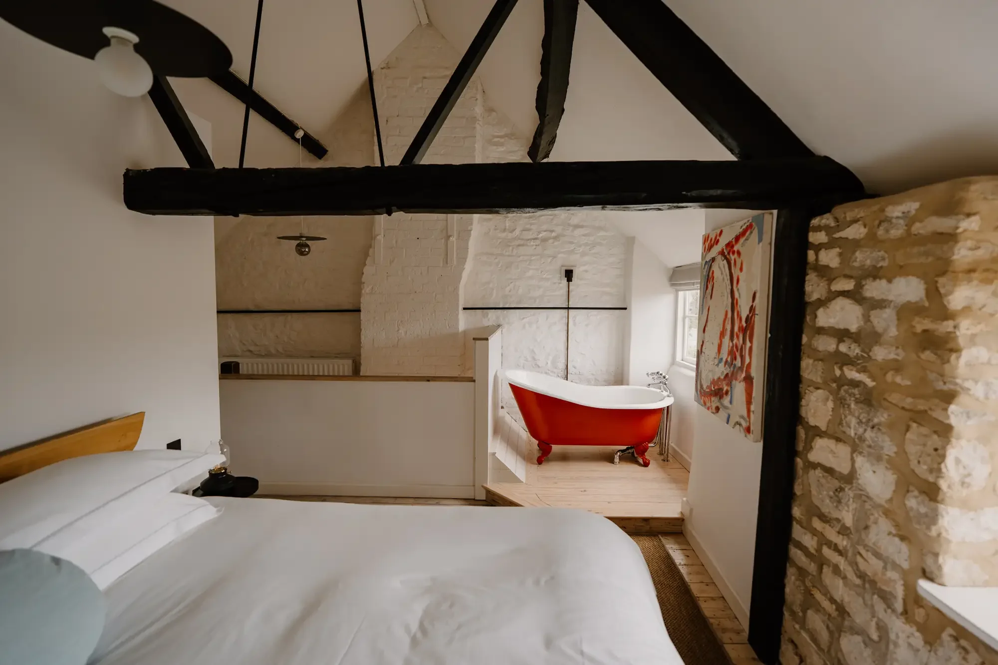 Interior view of a cozy room with white brick walls, a window, a red vintage-style bathtub, a colorful abstract rug, and exposed wooden beams.