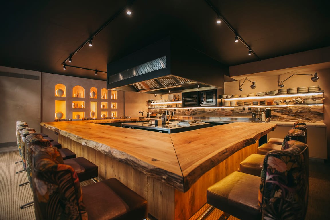 Interior view of a modern restaurant kitchen with a large wooden island at the center, surrounded by bar stools with patterned upholstery, decorative shelving on the walls, and a black range hood overhead.