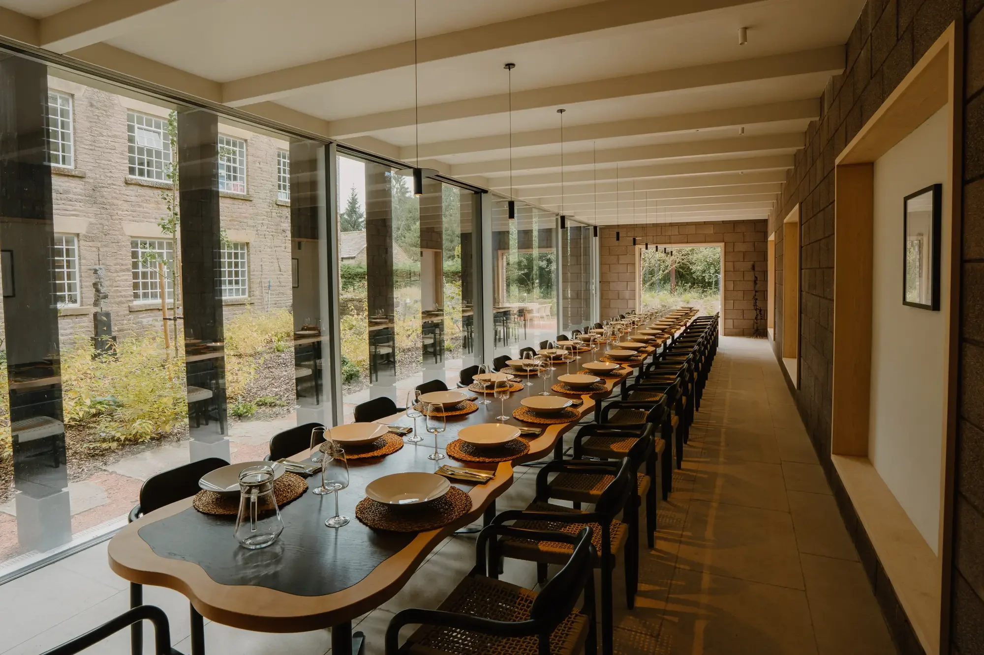 Long indoor dining table set with plates, glasses, and woven placemats, in a modern space with large windows and outdoor views.