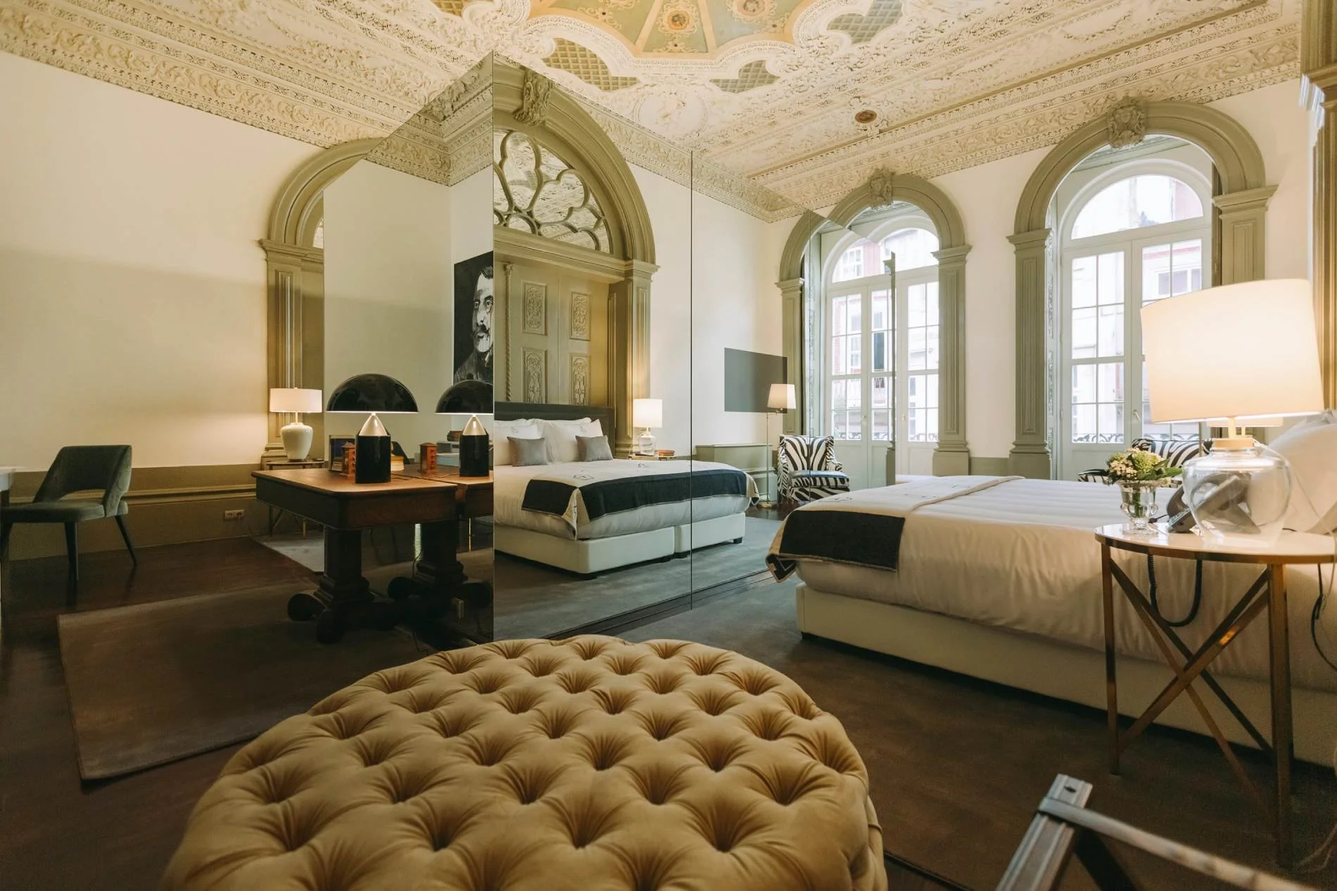 A luxurious hotel room with ornate ceiling details, large windows, and stylish furnishings including two beds, a mirrored wall, lamps, and decorative elements.