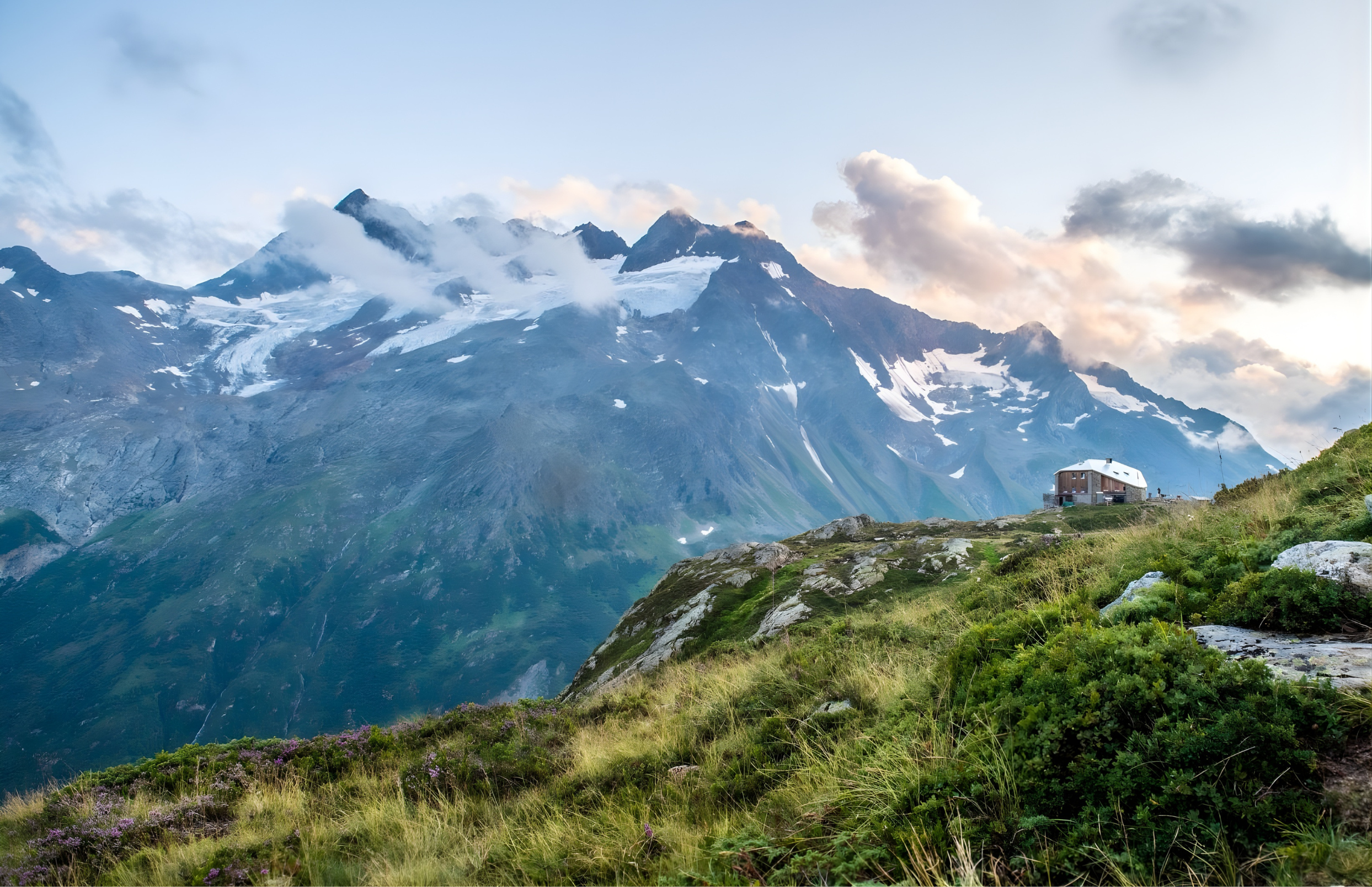 A mountain landscape with snow-capped peaks, clouds in the sky, and a small house on a green hillside.