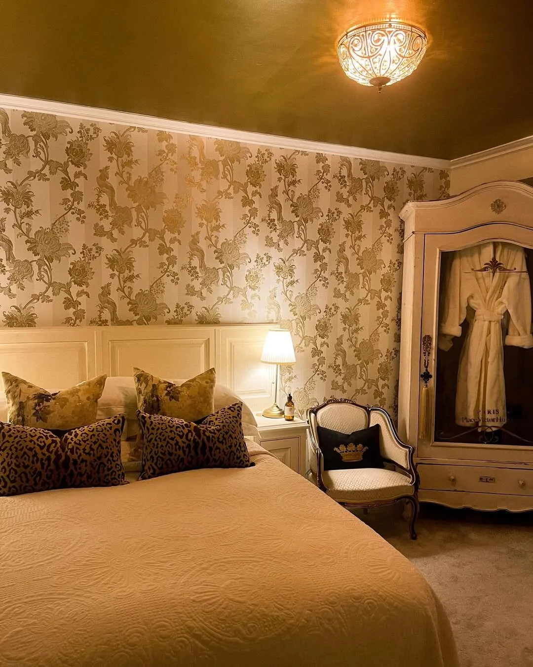 A luxurious bedroom with floral wallpaper, a bed with multiple decorative pillows, a bedside lamp, an elegant armchair with a crown-embroidered pillow, and a large wardrobe with curtains.