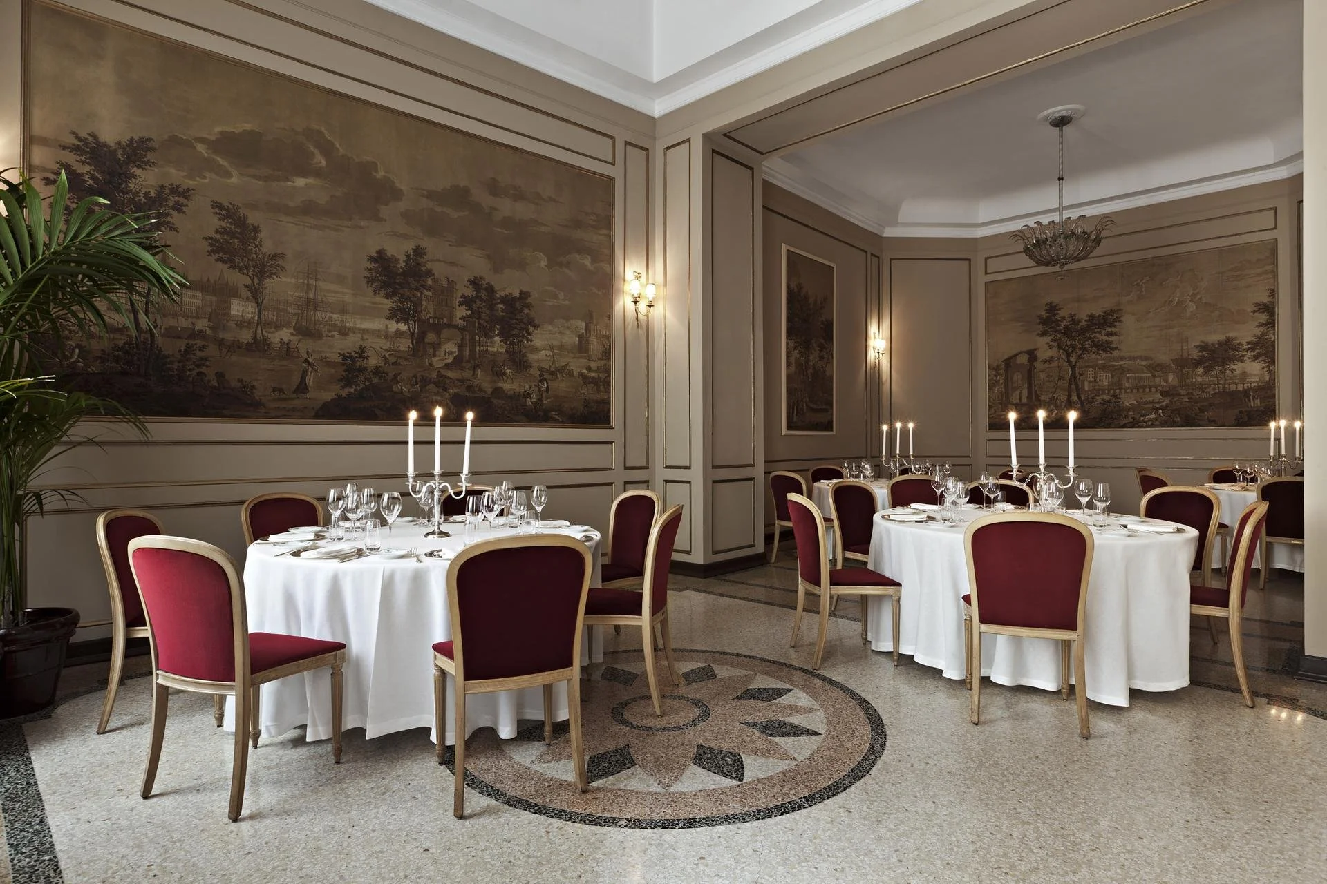 Elegant dining room with round tables covered in white tablecloths, set for a formal meal with wine glasses and silverware. Red upholstered chairs surround each table. The room features ornate wall moldings, large wall paintings depicting historical landscapes, and decorative wall sconces with lights. A chandelier hangs from the ceiling, and a potted plant is visible in the corner.