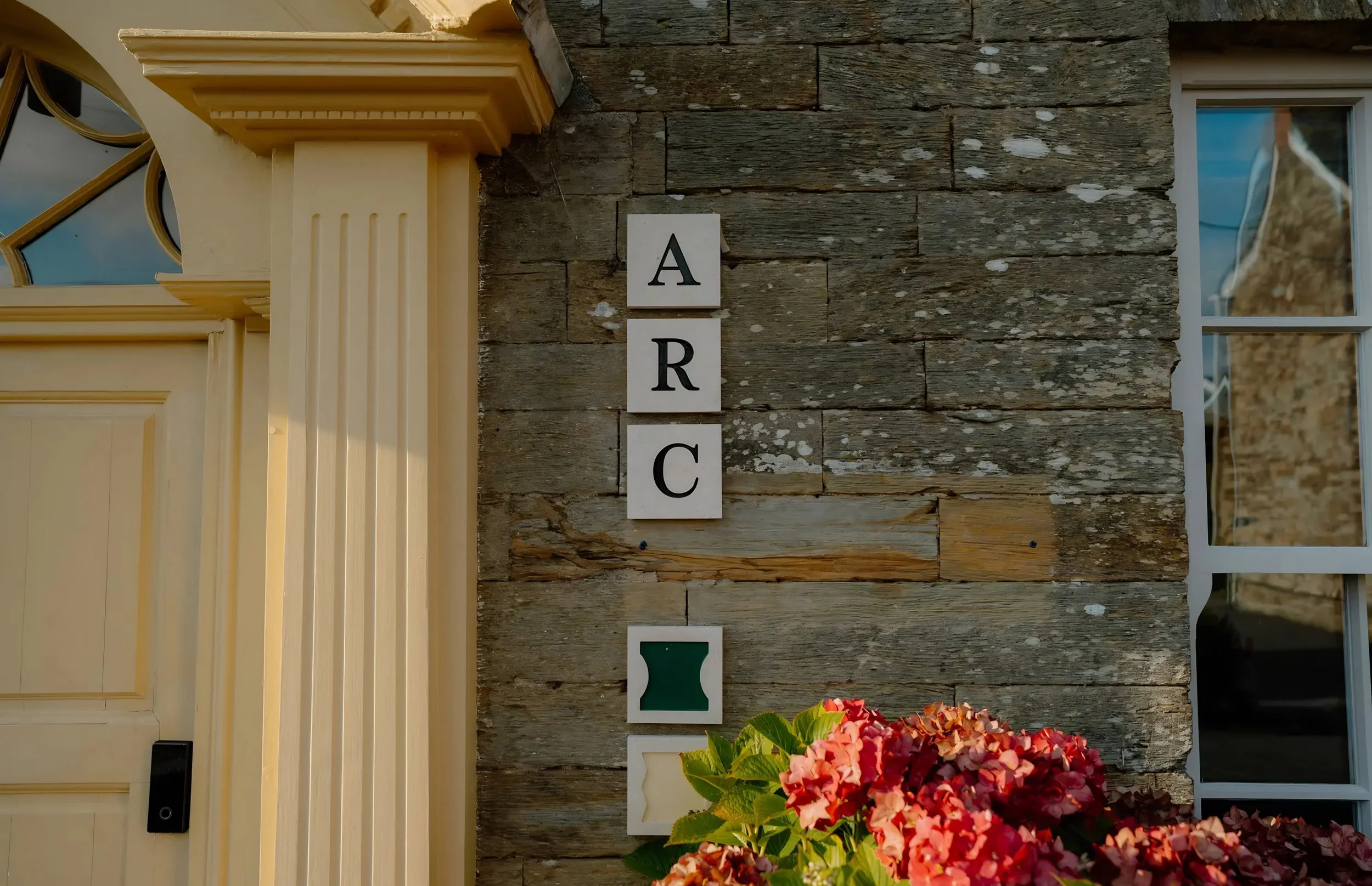 A house with a yellow door and trim, and a wooden exterior wall. There are vertical signs with the letters A, R, C, and E, and a house-shaped decoration. A red floral arrangement is in the foreground.