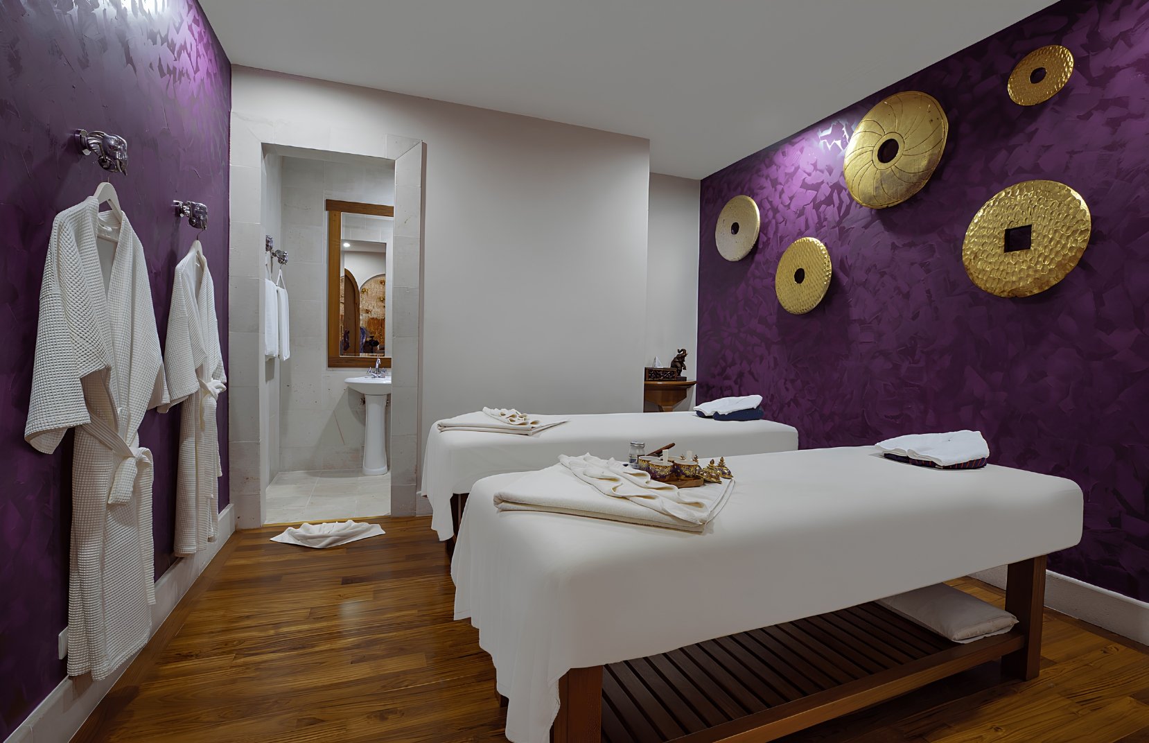 Spa room with purple textured accent wall decorated with large gold circular wall art, two massage tables with white linens, robes hanging on the wall, a small sink area visible through an open doorway, and wooden flooring.