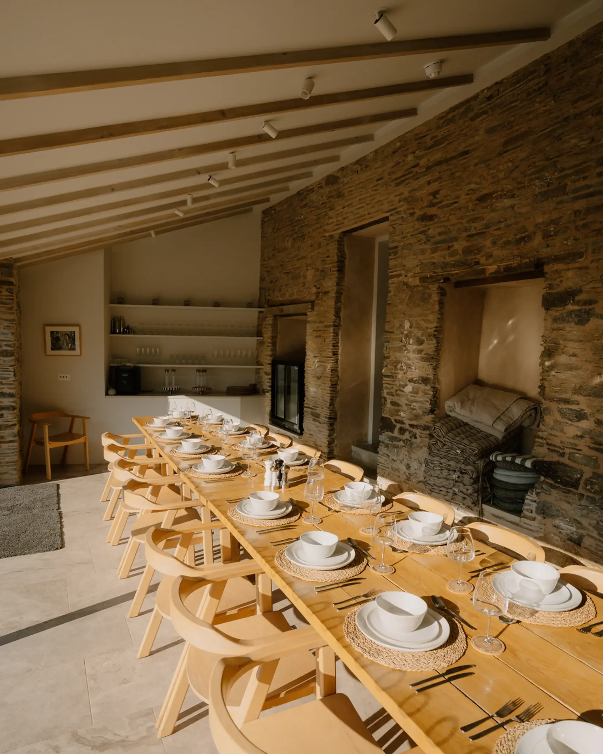 A dining area with a long wooden table set with white bowls, plates, wine glasses, and utensils. The table has woven placemats, and wooden chairs are arranged around it. The room features a rustic stone wall, beige tile flooring, and a ceiling with exposed wooden beams and recessed lighting.