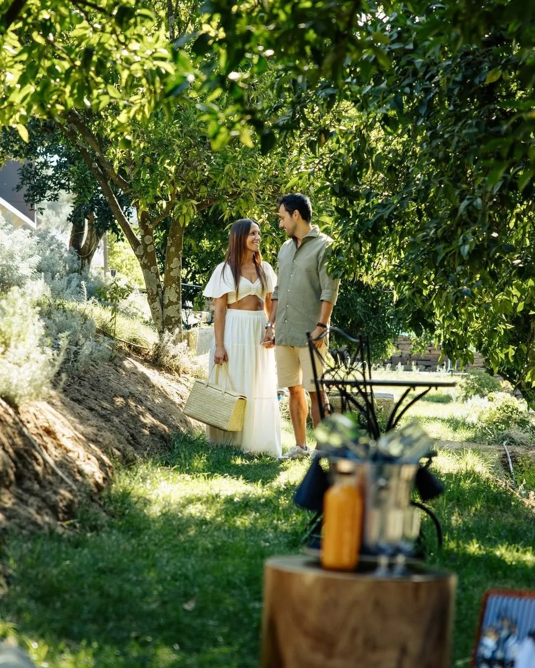 A man and woman standing together outdoors under green trees, smiling and holding hands, with a picnic setup in the foreground.