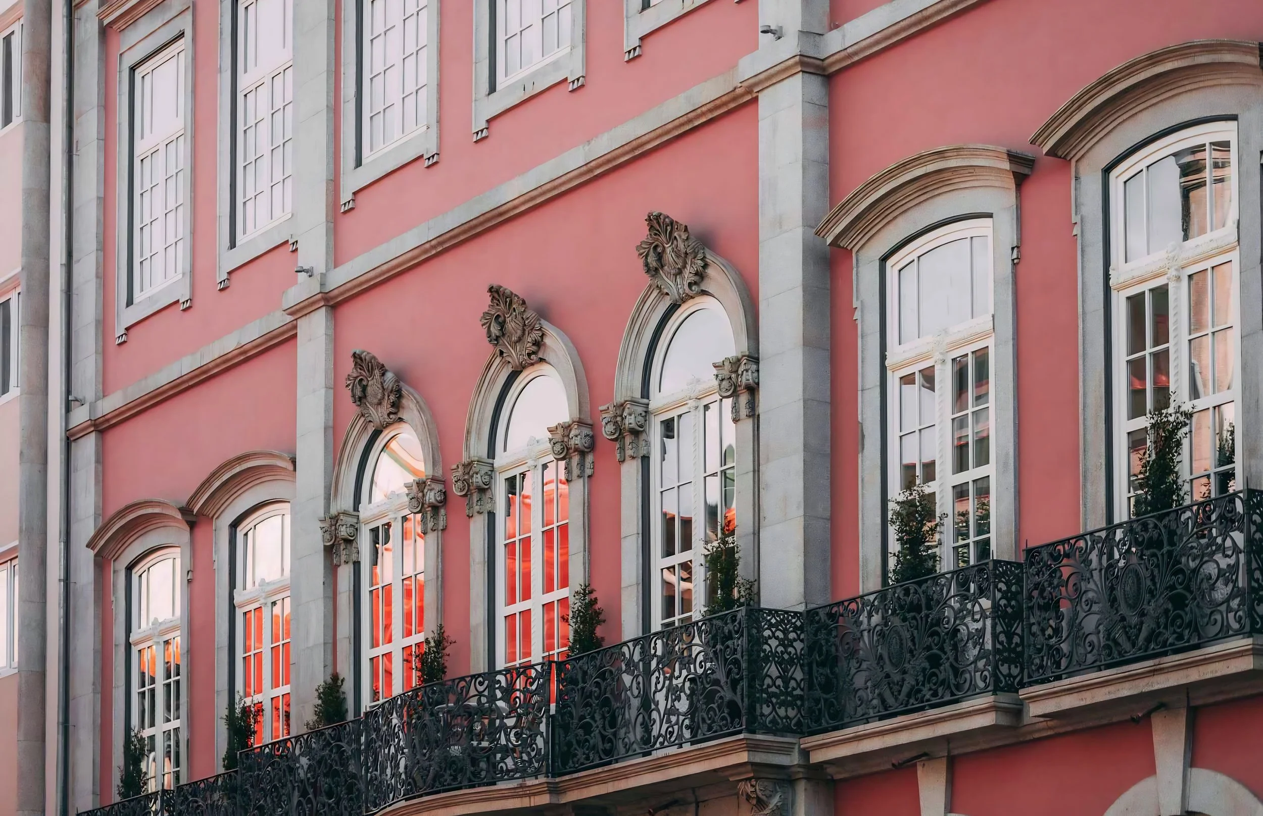 A pink building with large windows, ornate architectural details, and decorative lion head sculptures above some windows. There is a black wrought iron balcony with decorative patterns along the bottom of the building.