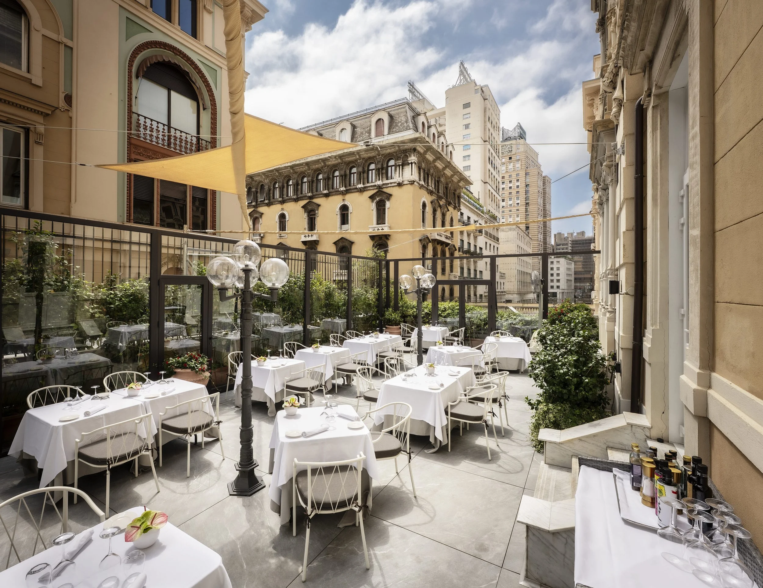 An outdoor dining area with round tables draped in white tablecloths, set with silverware and small floral centerpieces, enclosed by a black metal fence, with tall buildings in the background and a partly cloudy sky.