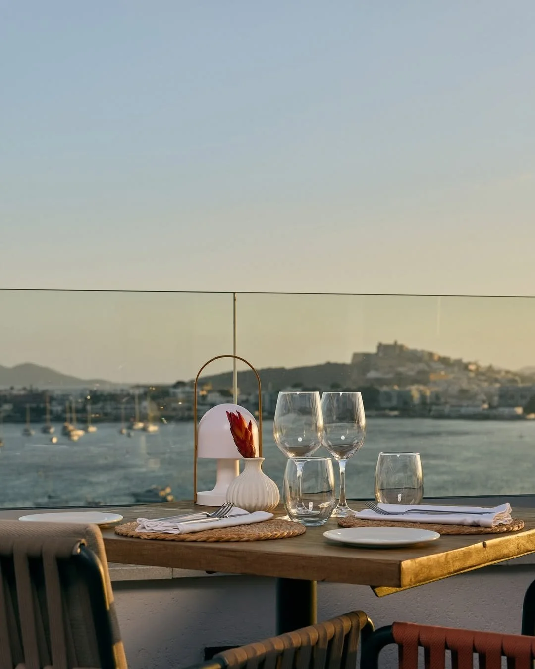 A table set for dinner with plates, glasses, and a small lamp, overlooking a body of water with boats and hills in the background during sunset.