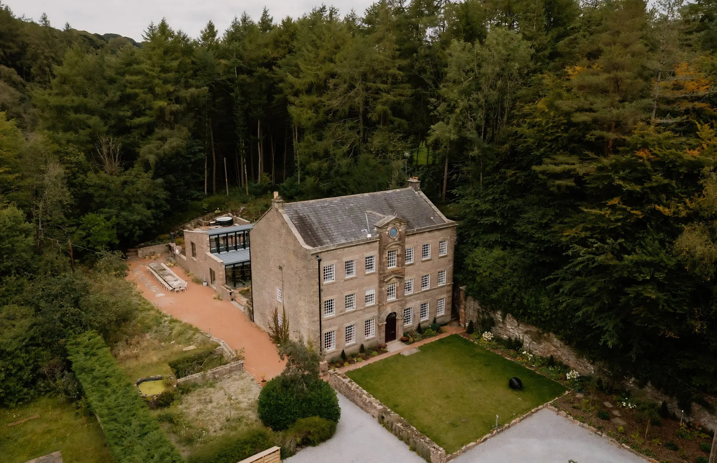 A large stone building with multiple floors and rows of windows, surrounded by lush green trees and a landscaped garden with a gravel pathway.
