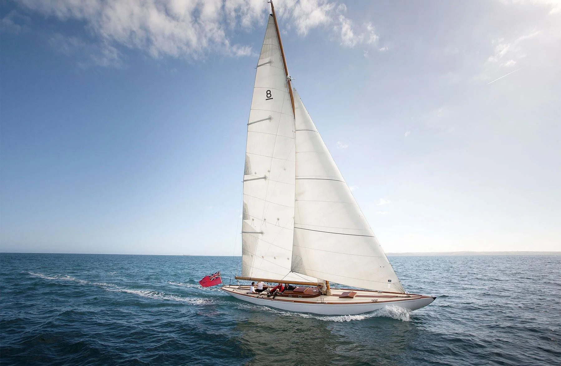 A sailboat with a white sail and a wooden hull sailing on open water under a partly cloudy sky, with a red flag at the stern and a small group of people on board.