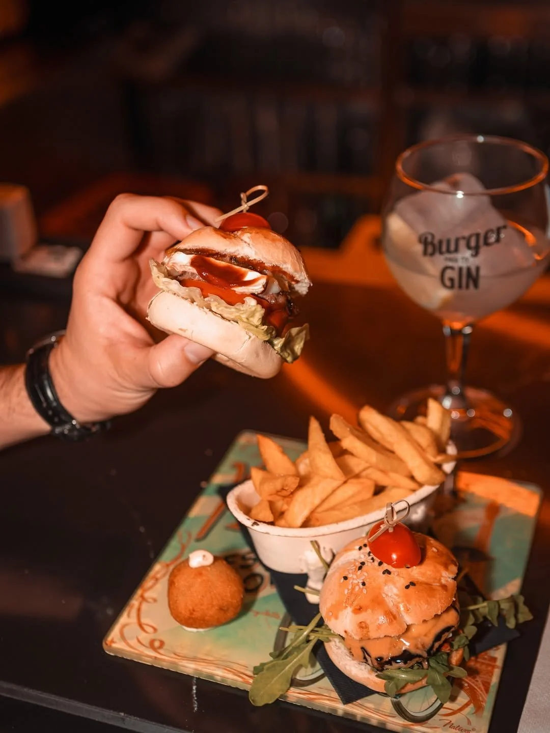 Hand holding a mini burger with ketchup, lettuce, and tomato. On the tray, there is another mini burger with tomato, a serving of French fries, and a fried appetizer. A decorative glass with ice and the words 'Burger GIN' is in the background.