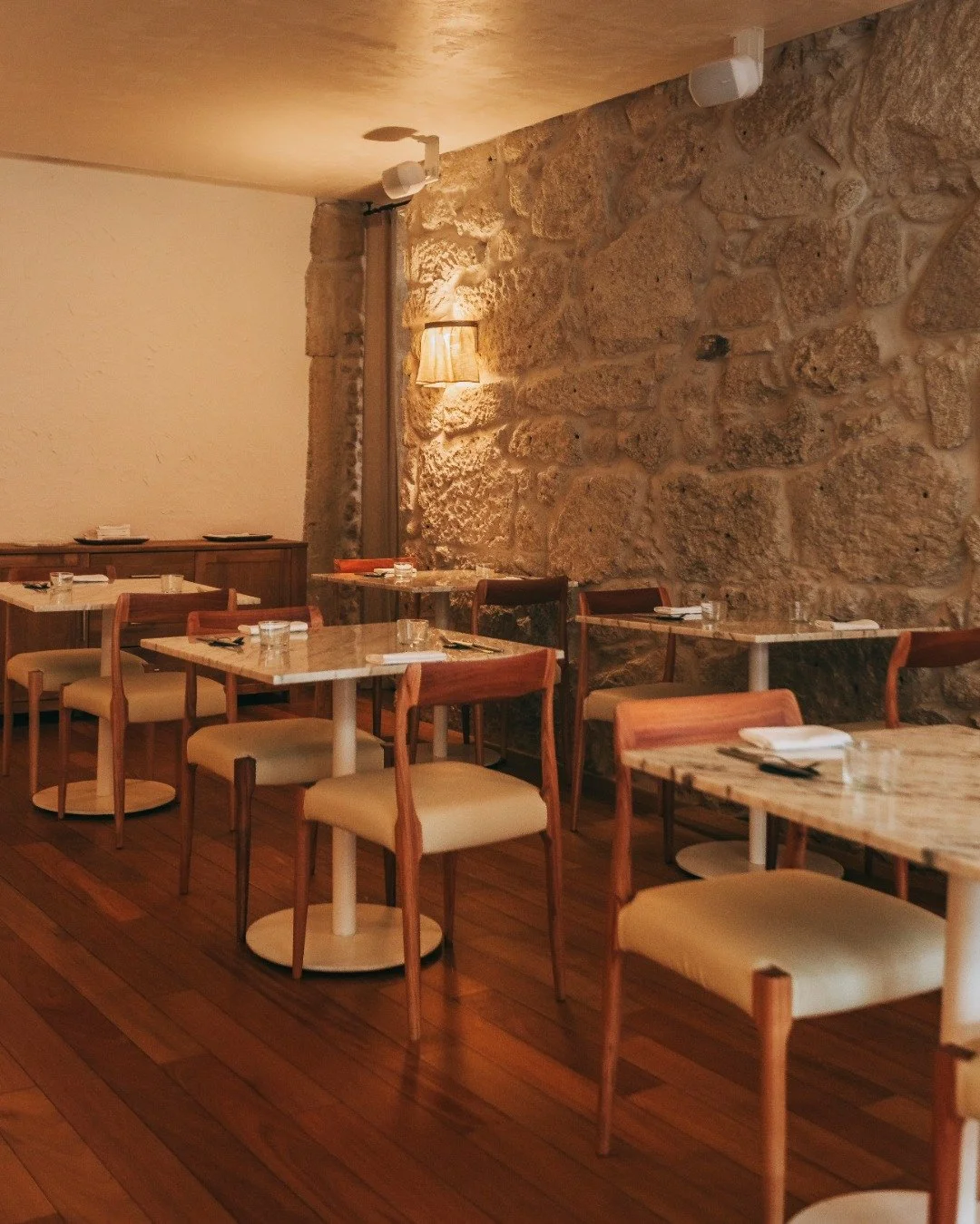 An empty dining area with marble tables, wooden chairs with cream cushions, a stone wall, a wooden floor, a wall-mounted lamp, and ceiling lights.