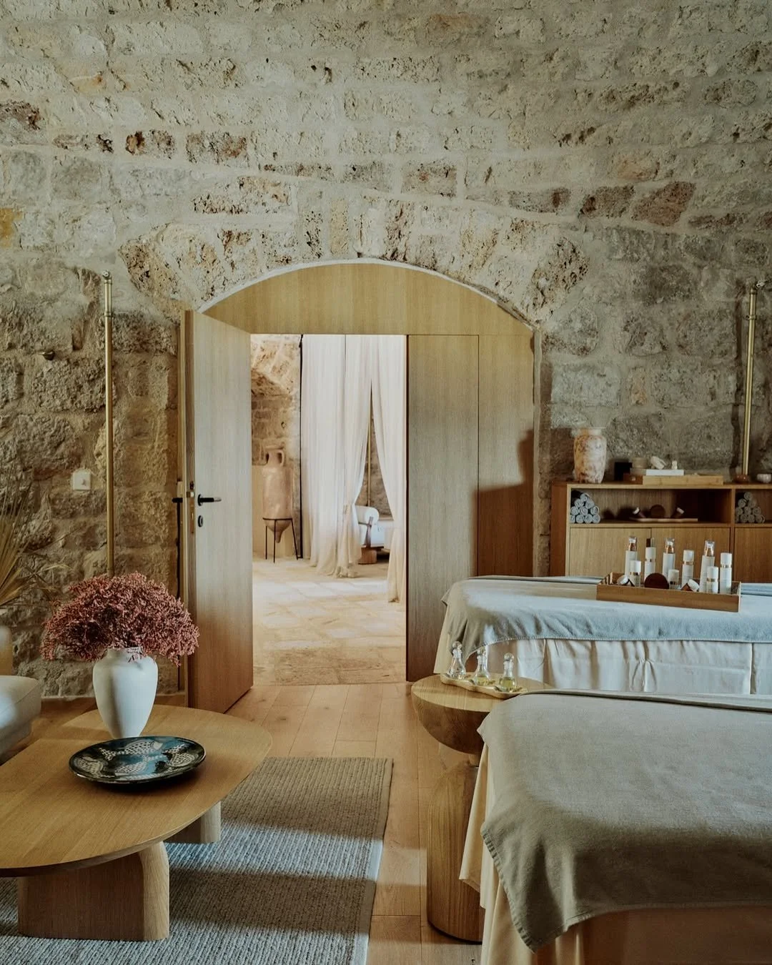 Interior of a rustic room with stone walls, wooden furniture, a vase with pink flowers on a low table, and a doorway leading to another room with sheer curtains and additional furnishings.