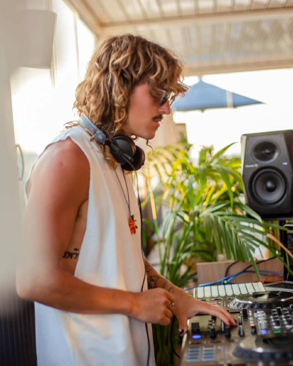 A person with curly hair, wearing sunglasses and headphones around their neck, operates DJ equipment outdoors near plants and a speaker.
