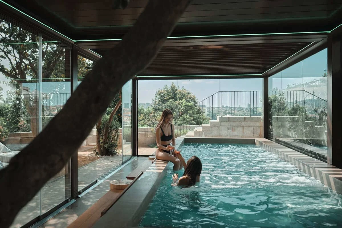 Two women enjoying a swim in an indoor pool with large glass windows showing outdoor greenery and a cityscape. One woman, sitting on the pool's edge, is pouring a drink, while the other is in the water.