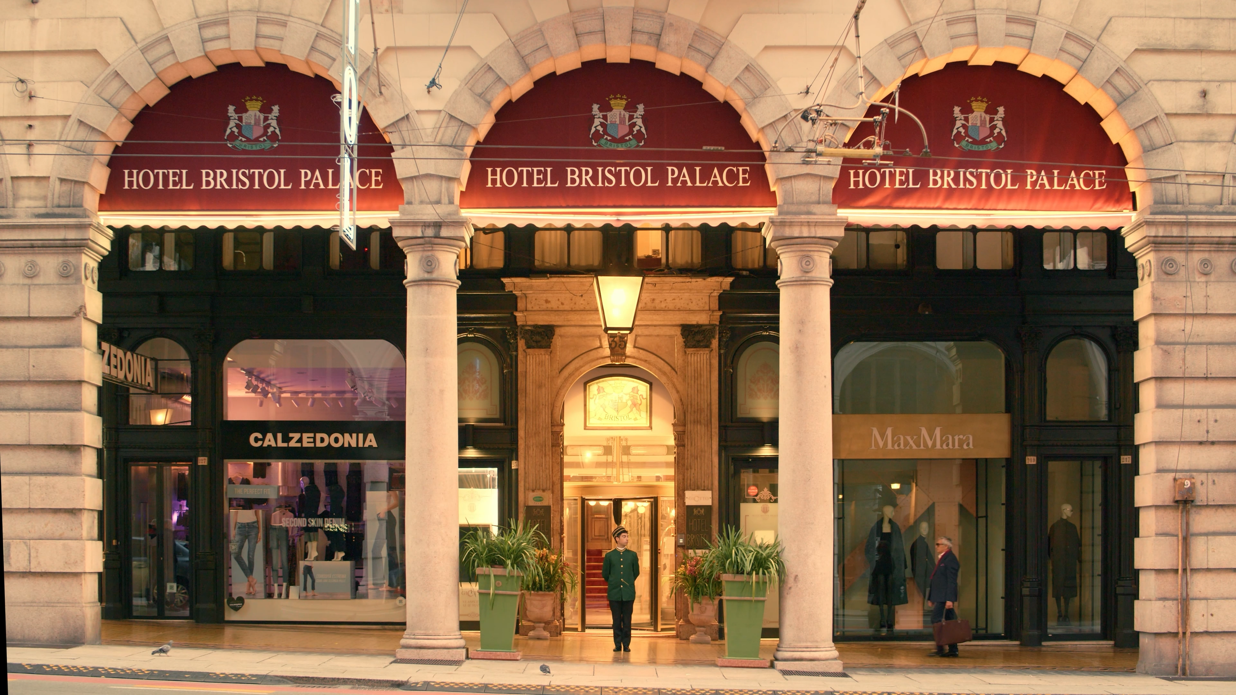 Facade of the Hotel Bristol Palace with a red awning displaying its name and coat of arms, columns at the entrance with people walking and standing outside, storefronts for Calzedonia and Max Mara on either side.