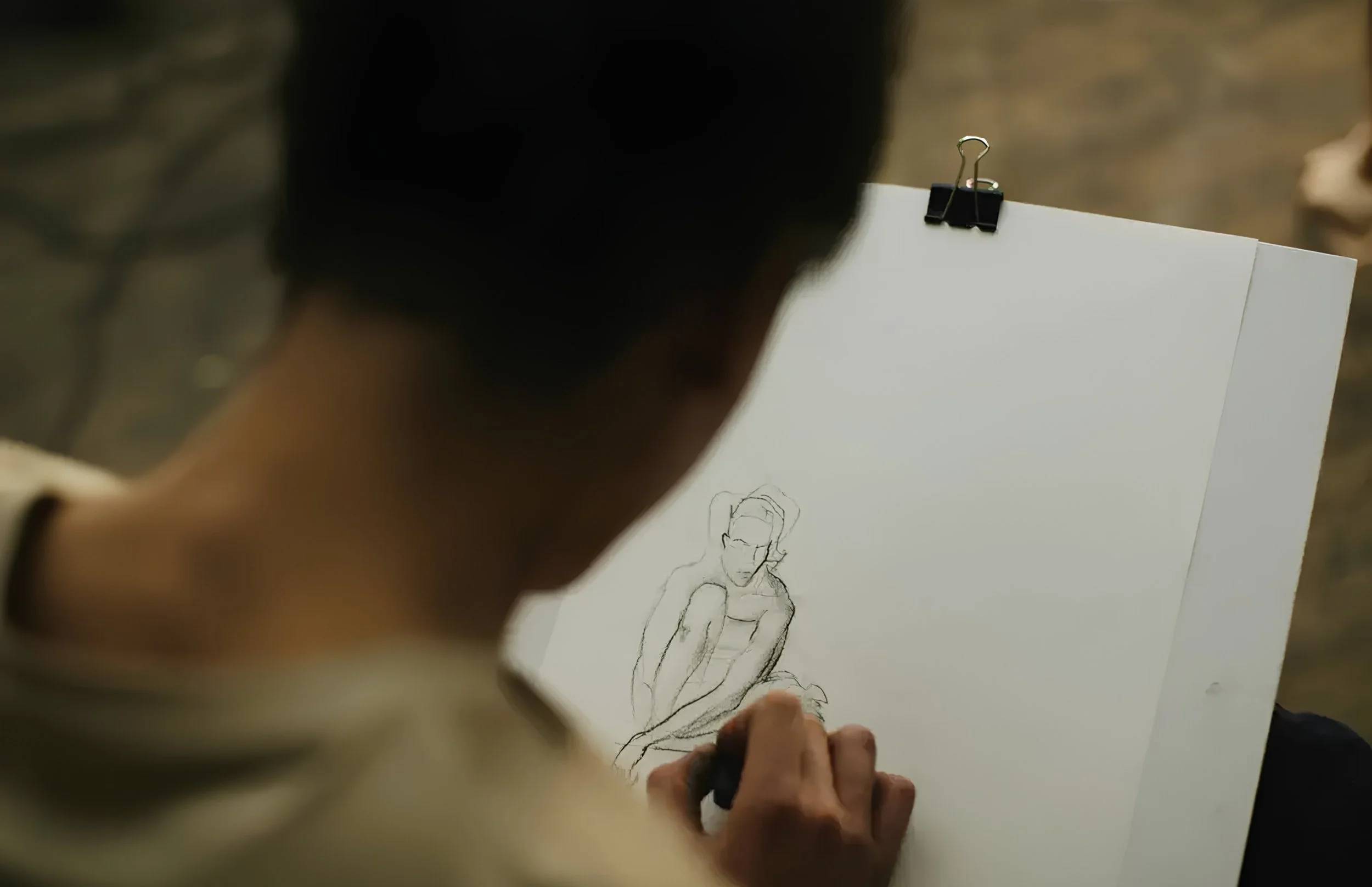 Close-up of a person sketching a figure on a sheet of paper, with the focus on the drawing and the person's hand holding a pencil.